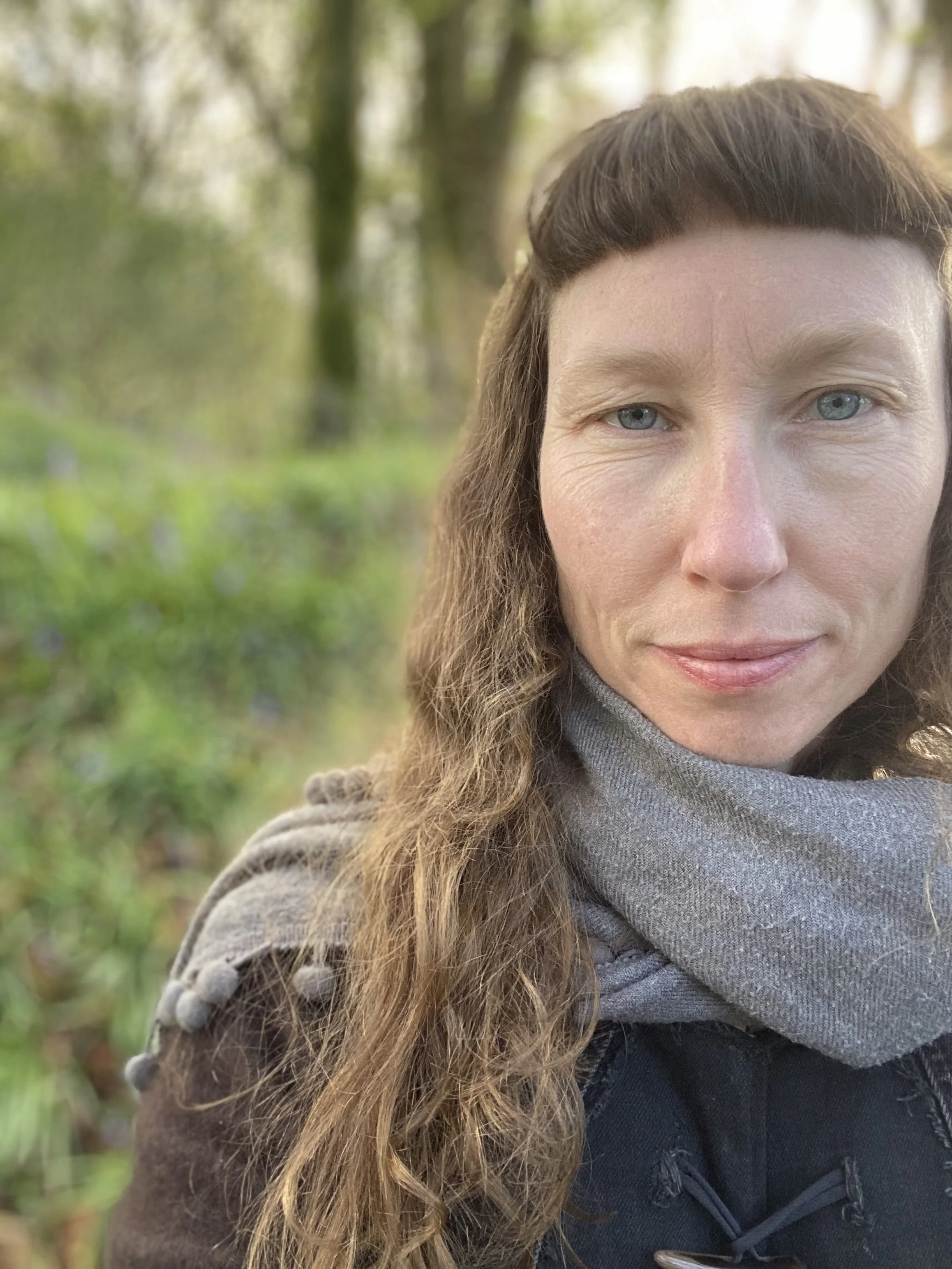 Gauri Algar with long, wavy hair, light skin, and blue eyes, wearing a gray hoodie and black jacket, outdoors in a green forested area with blurred trees in the background.