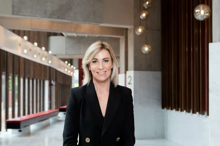 A woman with shoulder-length blonde hair, smiling, wearing a black blazer, standing in a modern indoor space with wooden accents and hanging spherical lights.