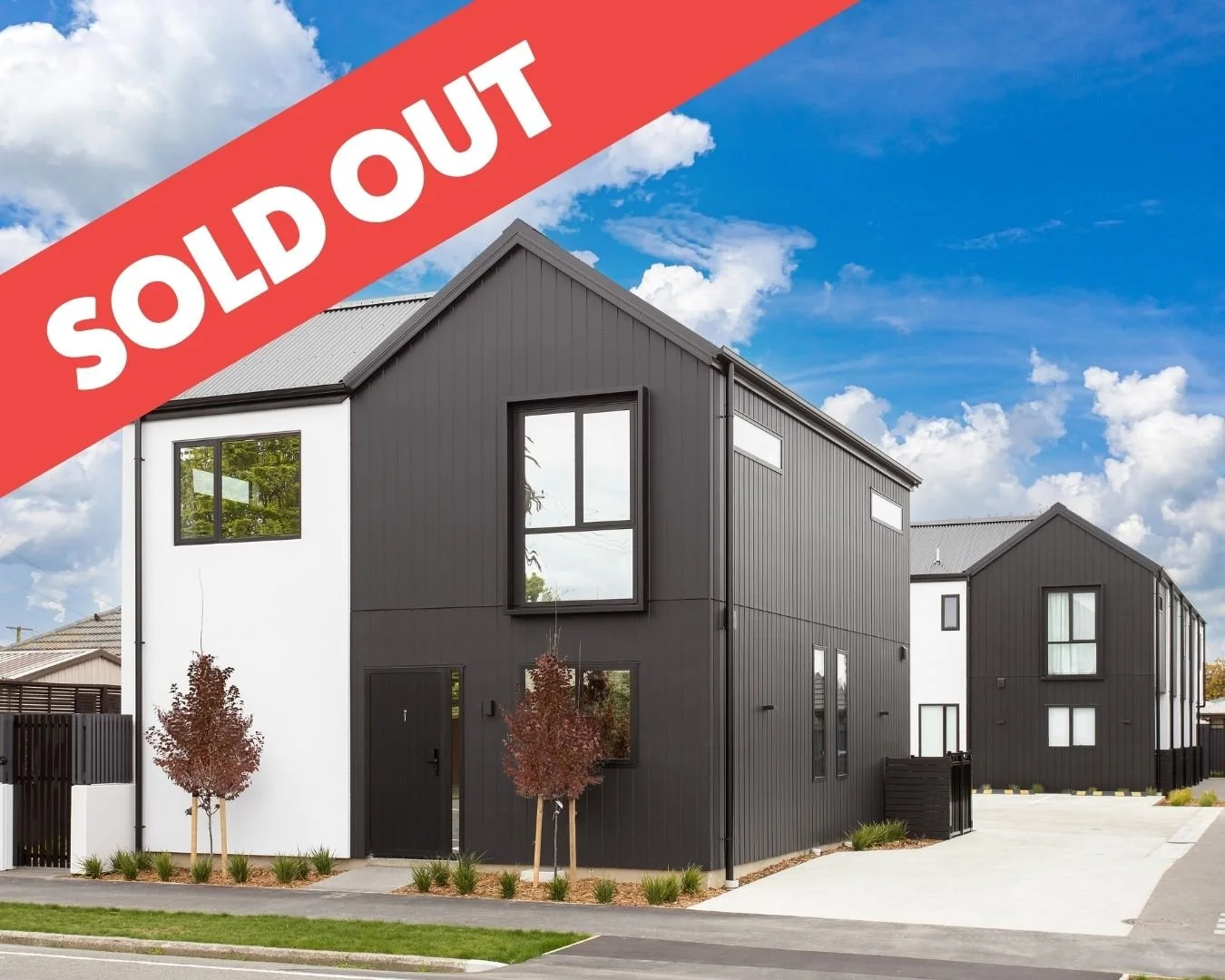 Modern black and gray residential buildings with landscaped front yards, pink flowering trees, and a car parked near the house. A red stripe with white text overlays the top left corner reading "SOLD OUT."