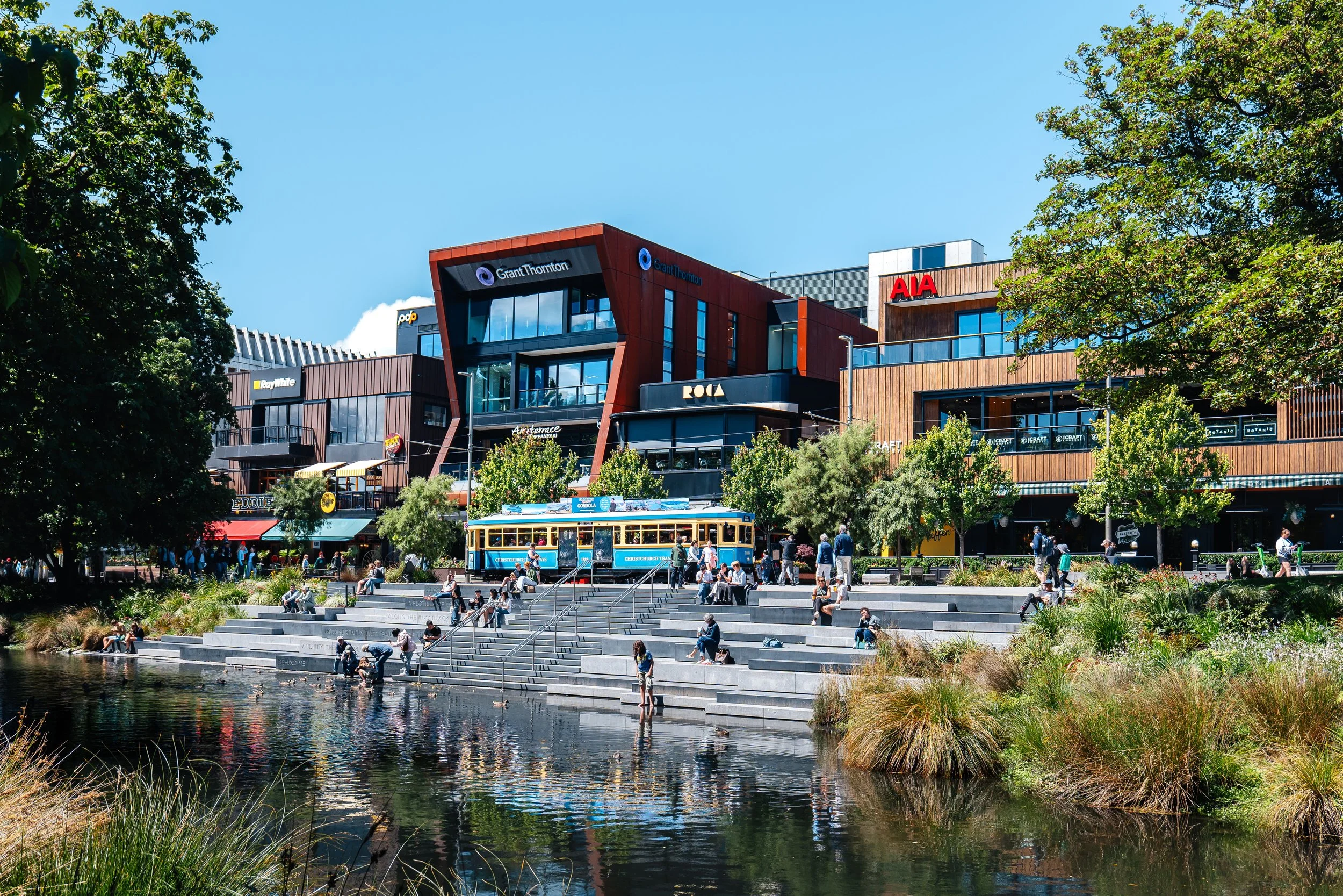 A bustling city scene featuring modern buildings with colorful signage, a tram, and a landscaped area with people sitting and walking by a water feature.
