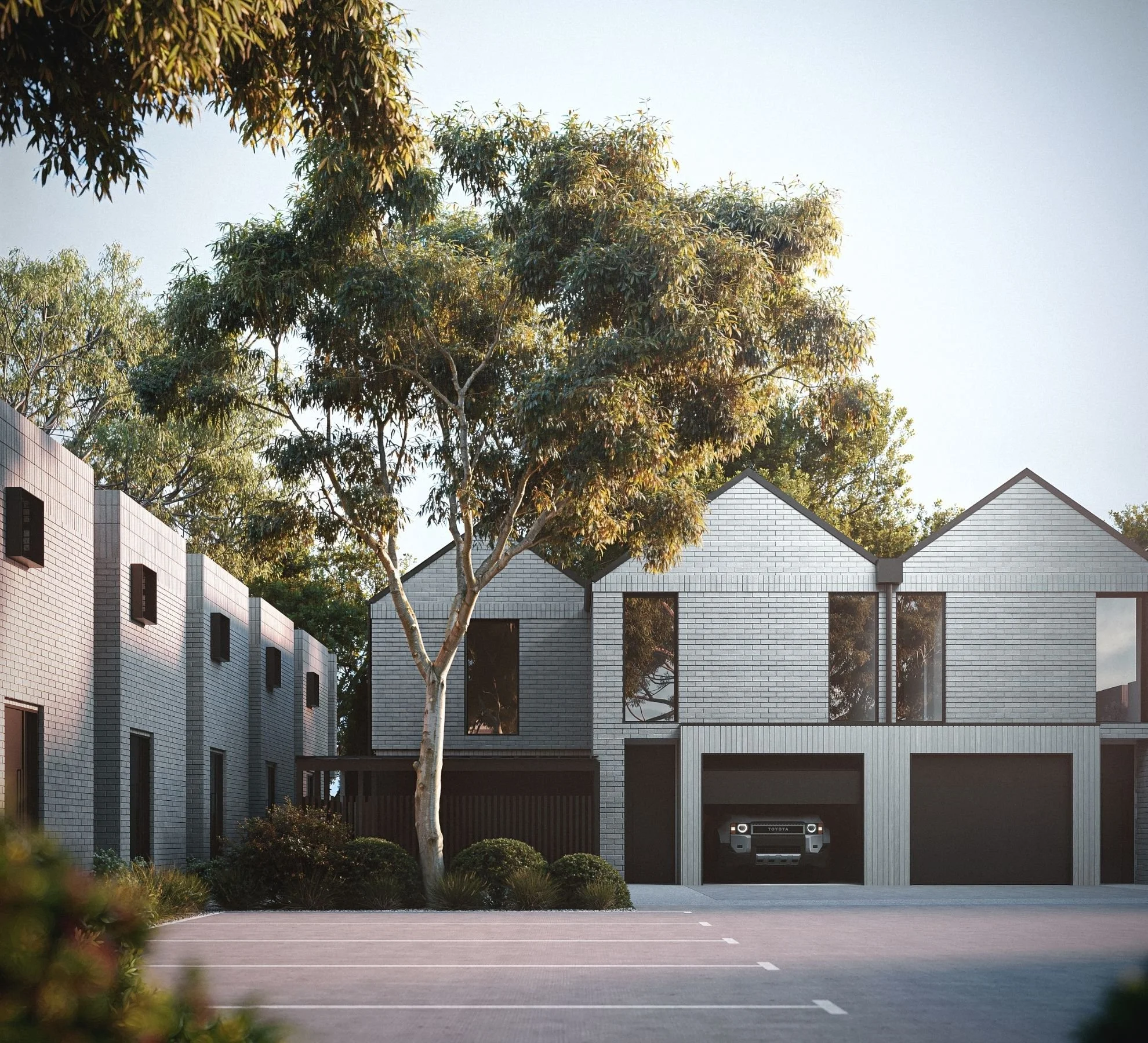 Modern residential housing with black brick buildings on the left, white brick houses with garage doors on the right, a large tree in the foreground, and a clear blue sky.