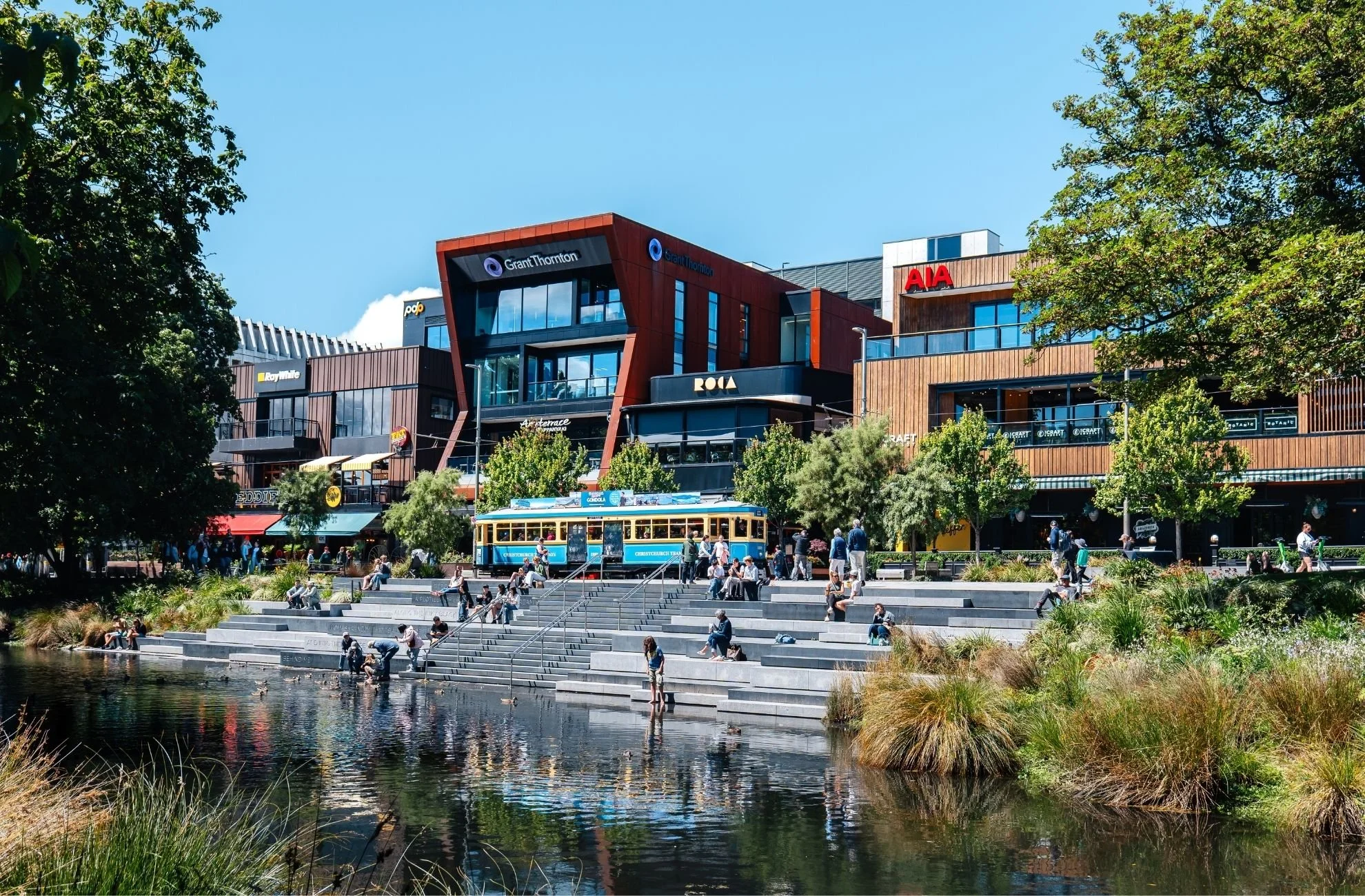 A bustling city scene featuring modern buildings with colorful signage, a tram, and a landscaped area with people sitting and walking by a water feature.