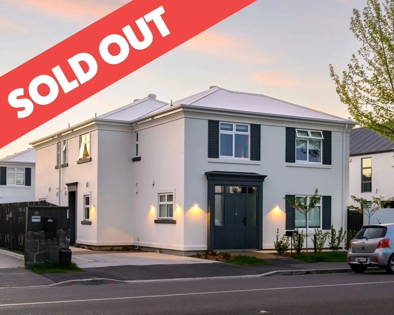 Modern white house with black shutters and front door, trees, parked car, and a sunset sky, marked sold out with a red banner across the top.