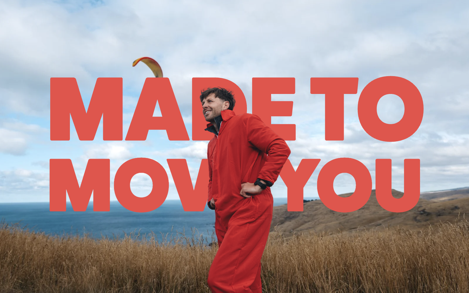 A man in a red jacket and red pants standing in a grassy field with a coastal landscape and cloudy sky in the background. The text "MADE TO MOVE YOU" is overlaid in large red letters.