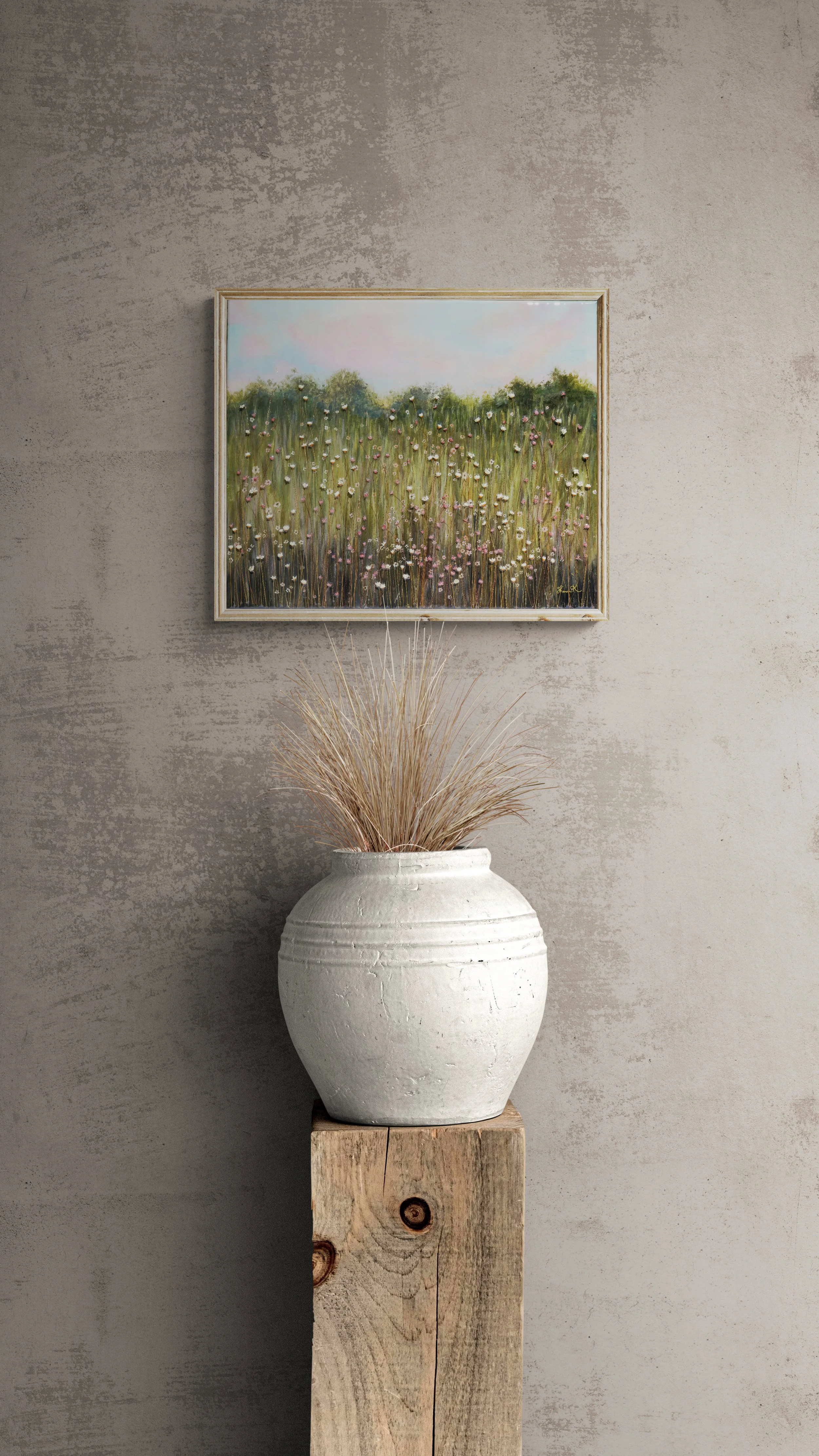 A white ceramic vase with dried grass sits on a wooden pedestal. Above it, a framed painting of a grassy field with flowers is hung on a textured concrete wall.