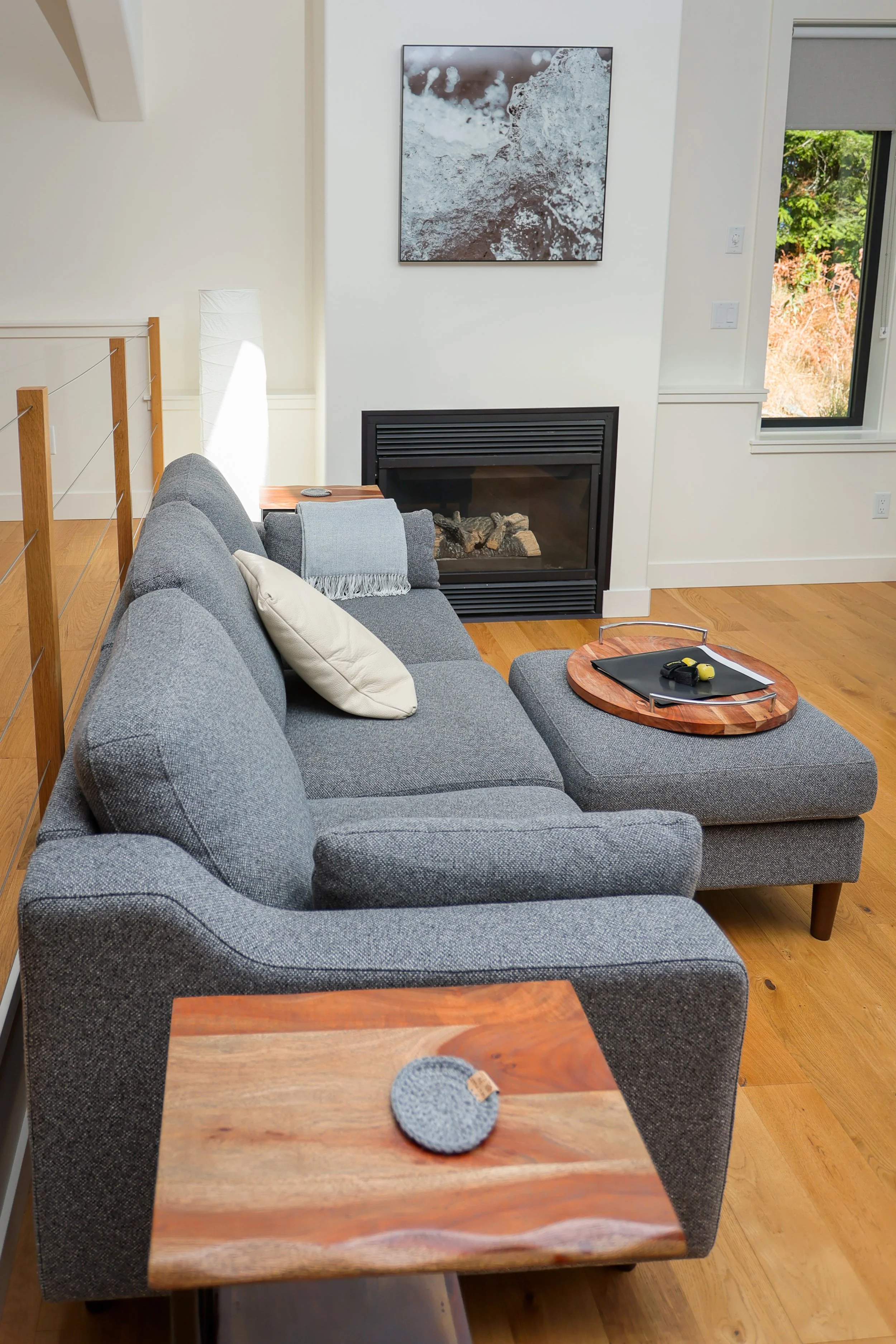 A living room with a gray fabric sectional sofa, light-colored throw pillows, a wooden coffee table with a tray and some accessories, a fireplace with a black frame, a white wall art piece, a window showing greenery outside, and a wooden floor.