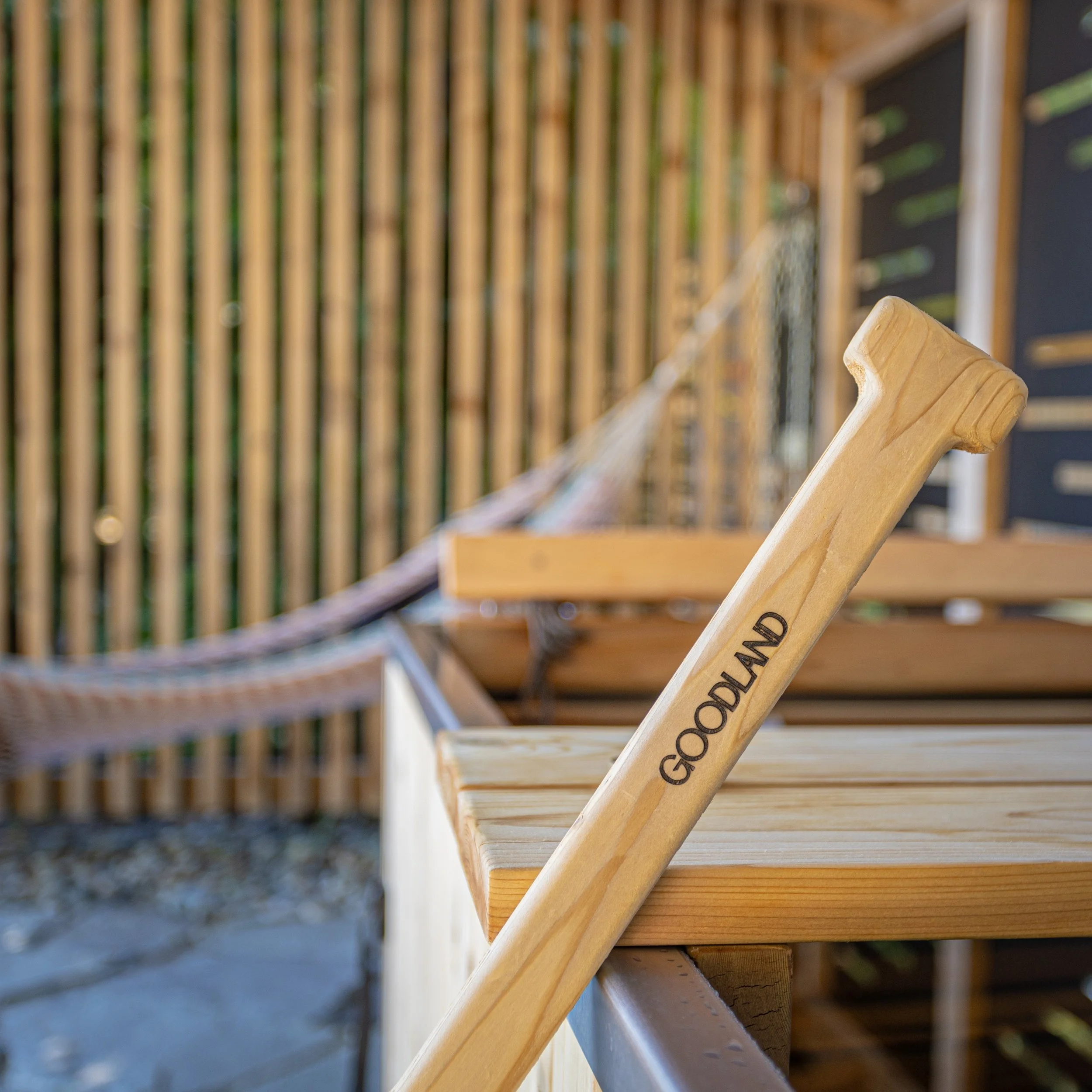 Close-up of a wooden croquet mallet with the word 'Goodland' engraved on it, resting on a wooden surface outdoors at a recreation area with a hammock and wooden fence in the background.