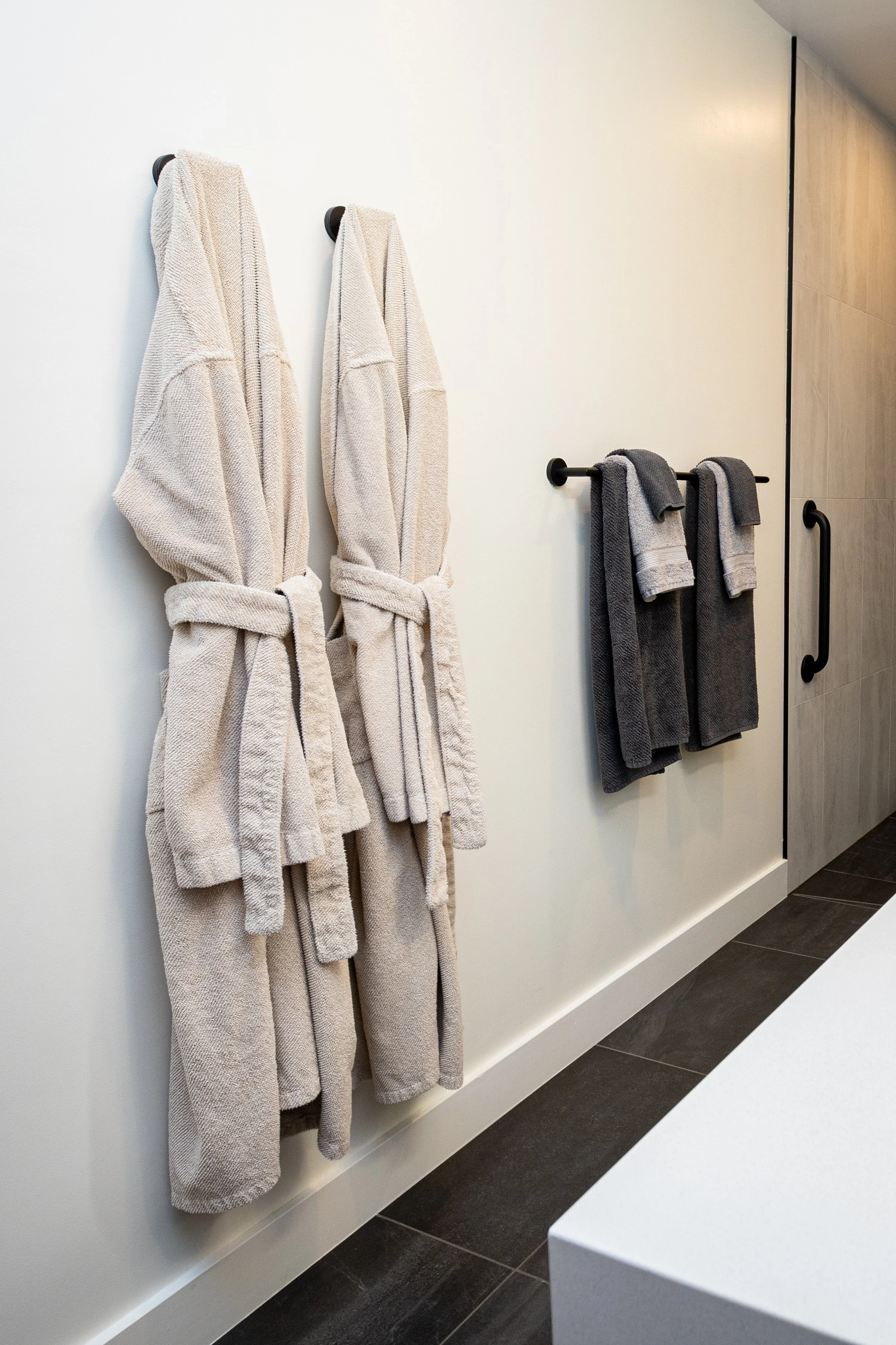 Two beige bathrobes hanging on hooks on a white wall, and two dark gray towels hanging on a rod nearby.