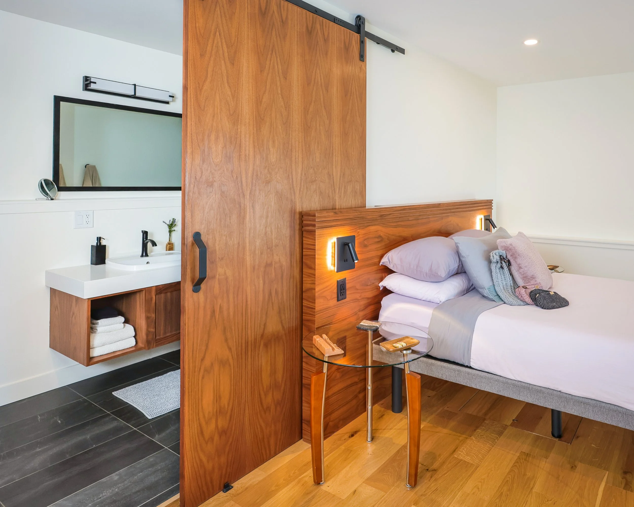 A bedroom with a wooden sliding door, a bed with pillows, a glass side table, and a small white sink with towels and a mirror in the background.