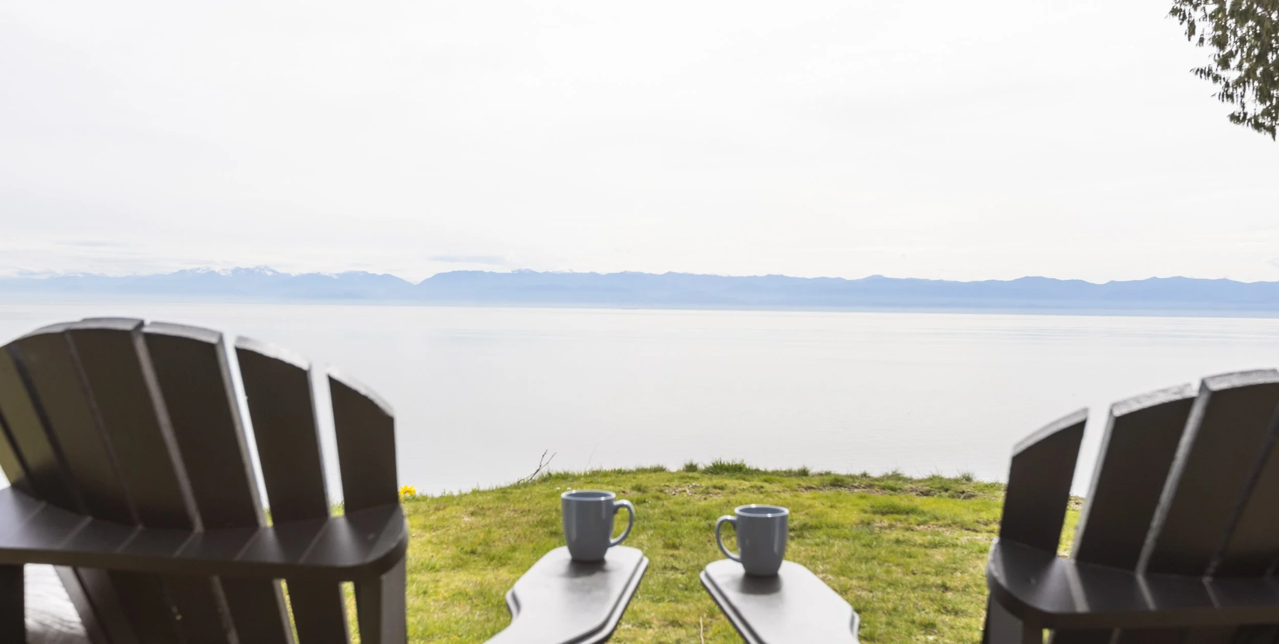 Two Adirondack chairs facing a scenic view of a lake with mountains in the distance, with two coffee mugs on the small table between them, on a grassy area.