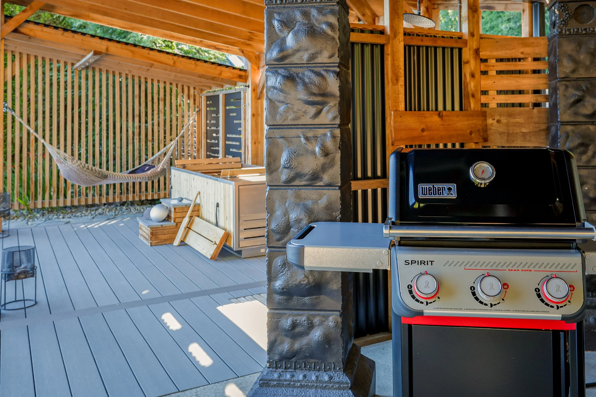 Outdoor patio with a black Weber grill in the foreground, wooden privacy fence, hammock, and outdoor kitchen area with wooden cabinetry and a stone pillar.