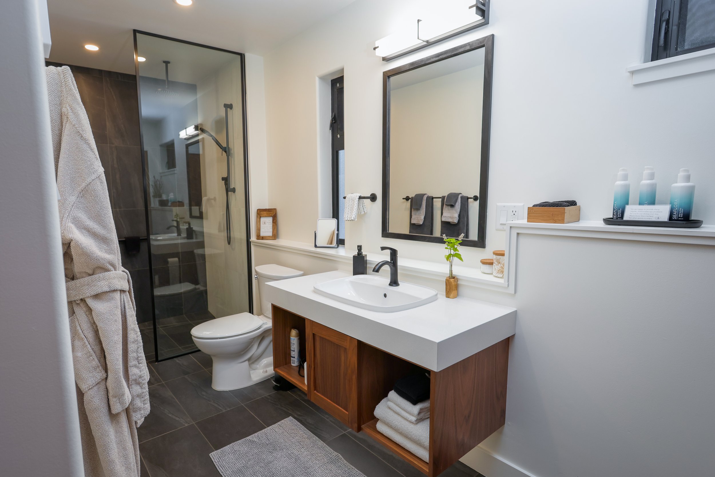 Modern bathroom with a glass shower stall, white toilet, wooden vanity with a white sink, large mirror, small window, and various toiletries and decor items.