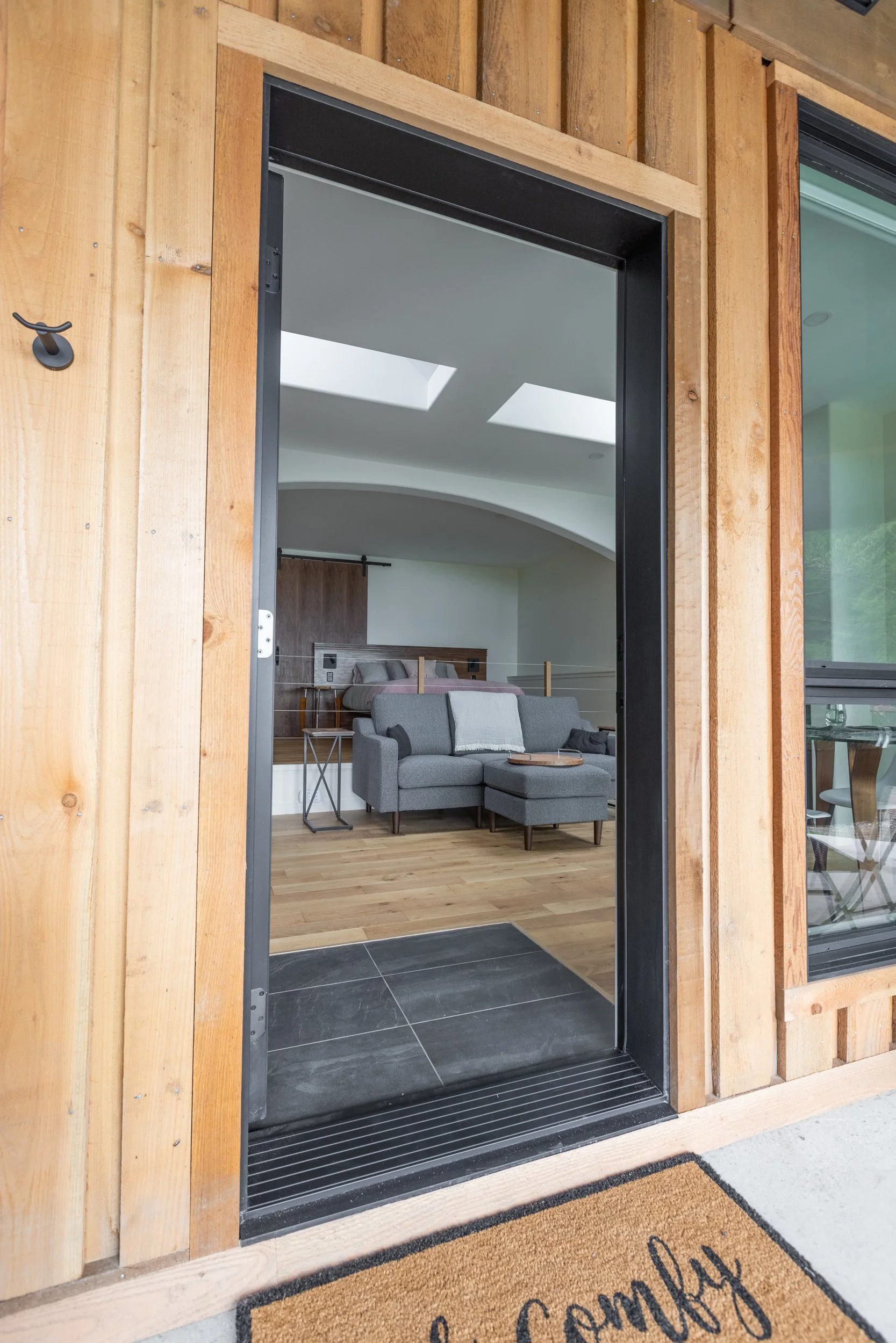 View through open doorway into a modern living room with a gray sectional sofa, wooden floors, and skylights.
