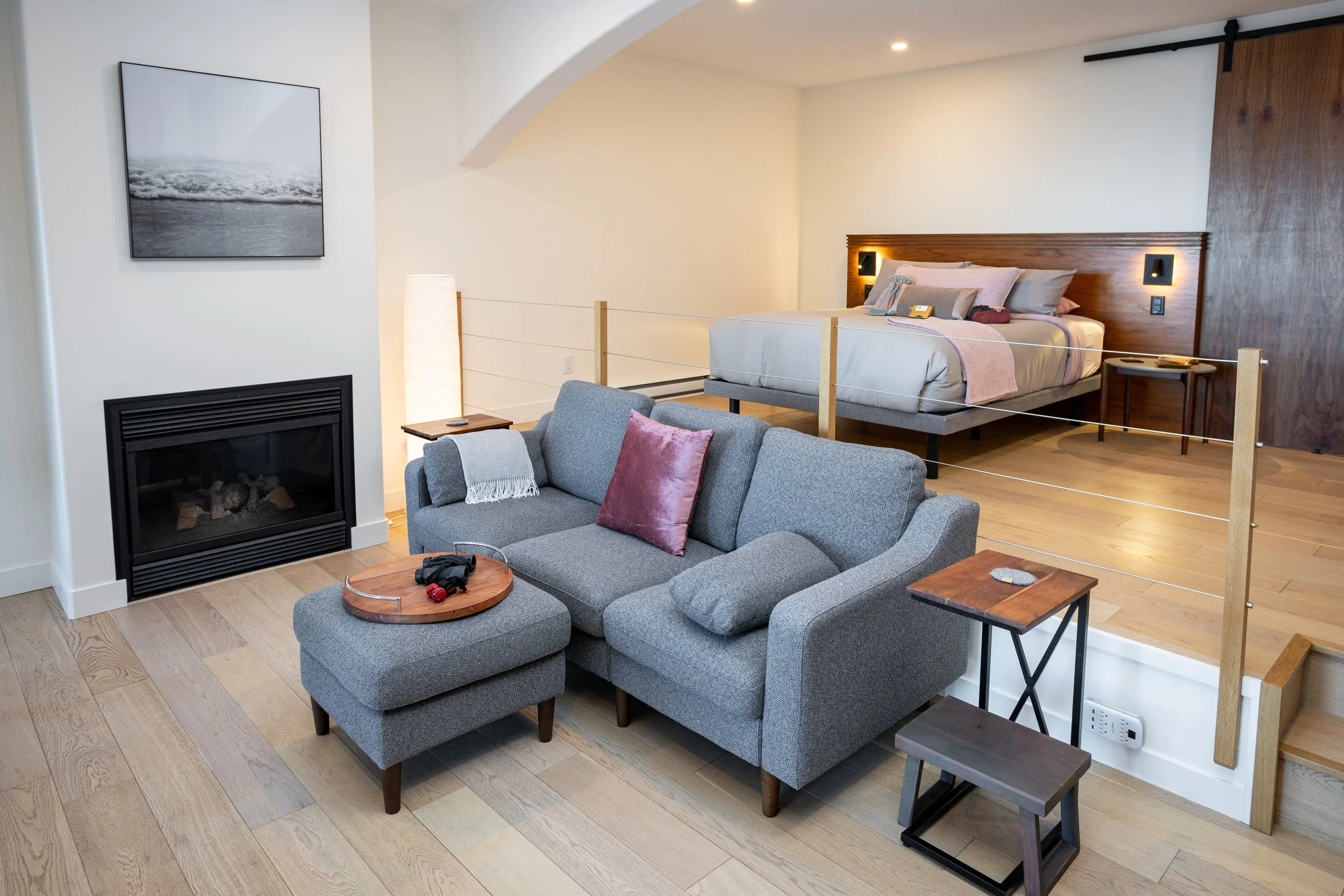 A modern living room with a gray sofa, pink cushions, and a wooden coffee table with headphones. A fireplace is on the left wall, with a framed photograph above it. In the background, a raised bedroom with a bed, nightstands, and wall-mounted lights is visible, separated by a wooden and rope barrier.