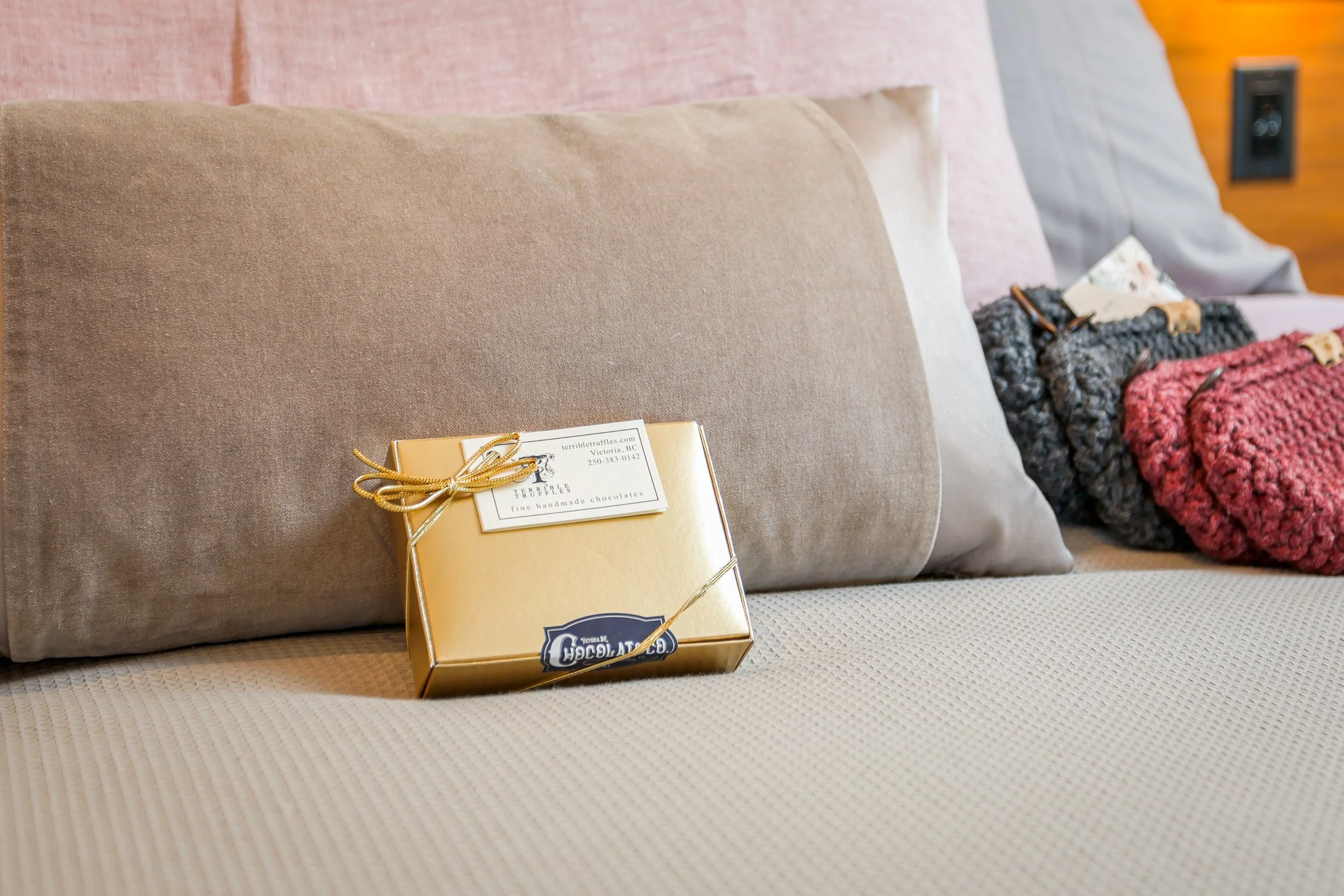 A small, gold-colored box of handmade chocolates from Terrible Truffles on a beige bedspread, with cushions and knitted blankets in the background.