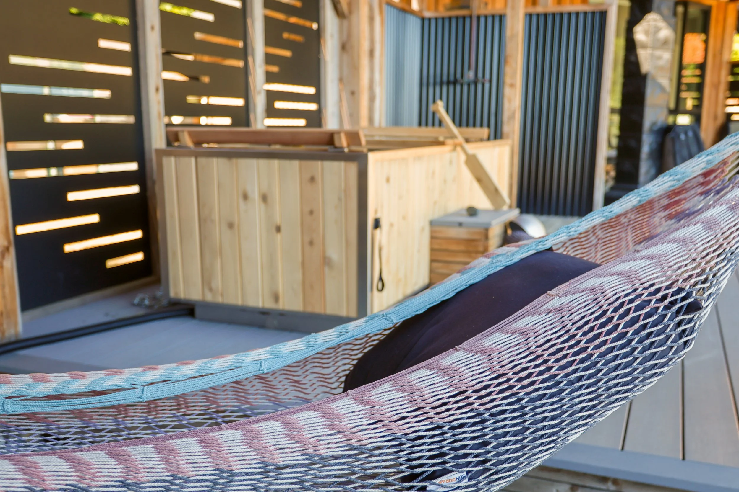 A hammock with a pillow hanging on a deck with a hot tub and wooden fencing in the background.