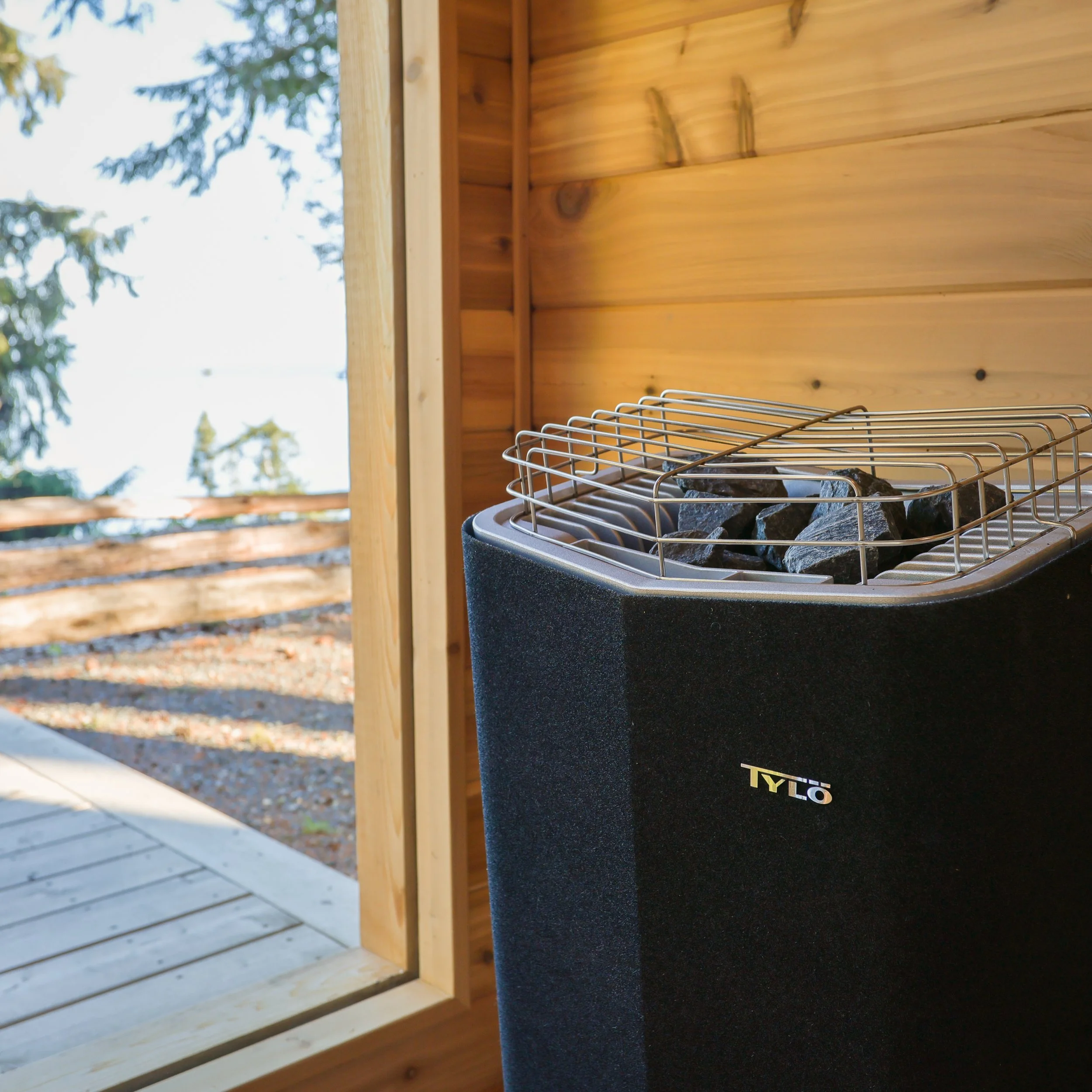 A sauna heater with stones inside, positioned inside a wooden sauna with an open window showing trees outside.