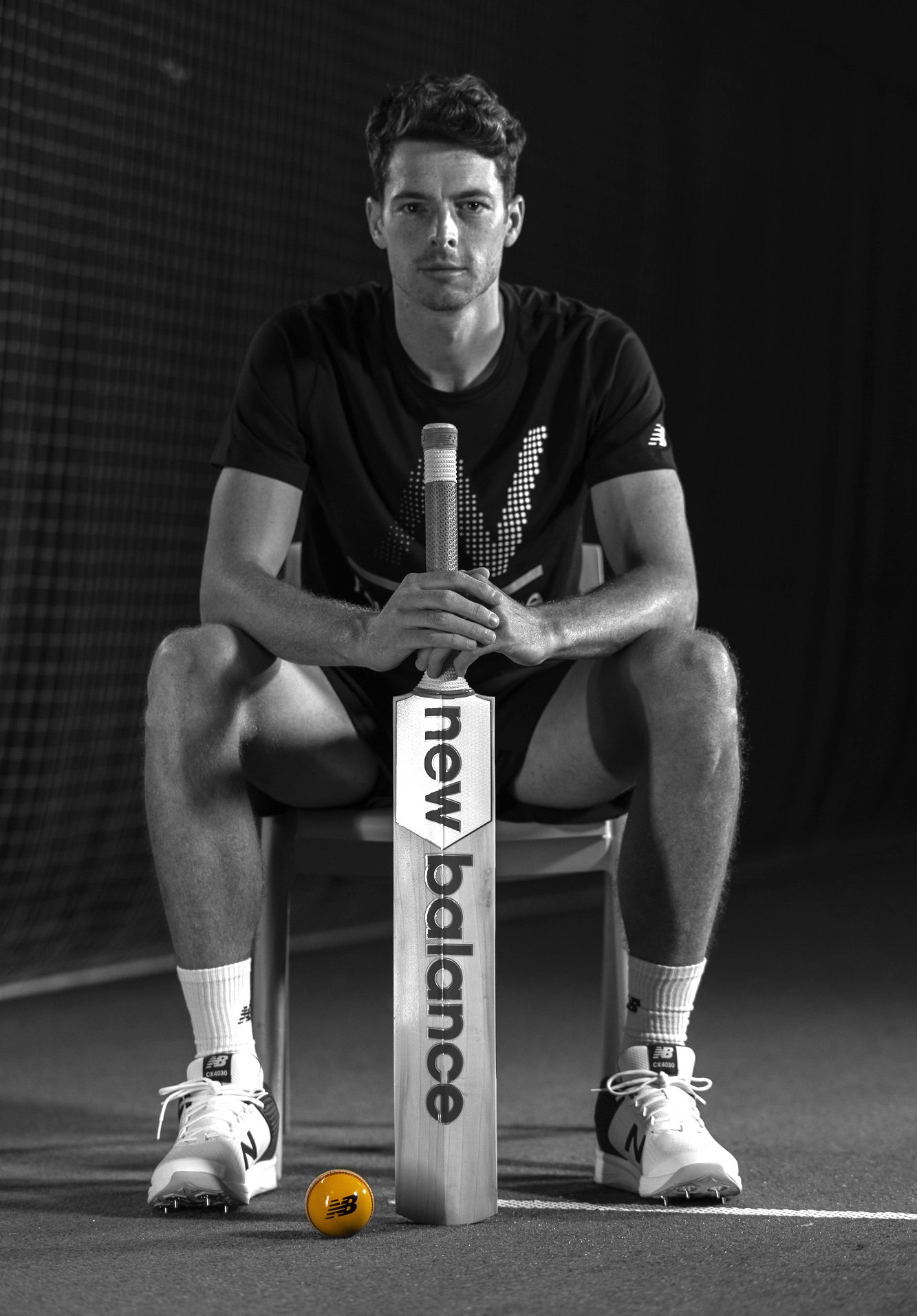 Person sitting on a chair with a cricket bat marked 'New Balance' resting on the ground, monochrome image with a yellow ball in front.