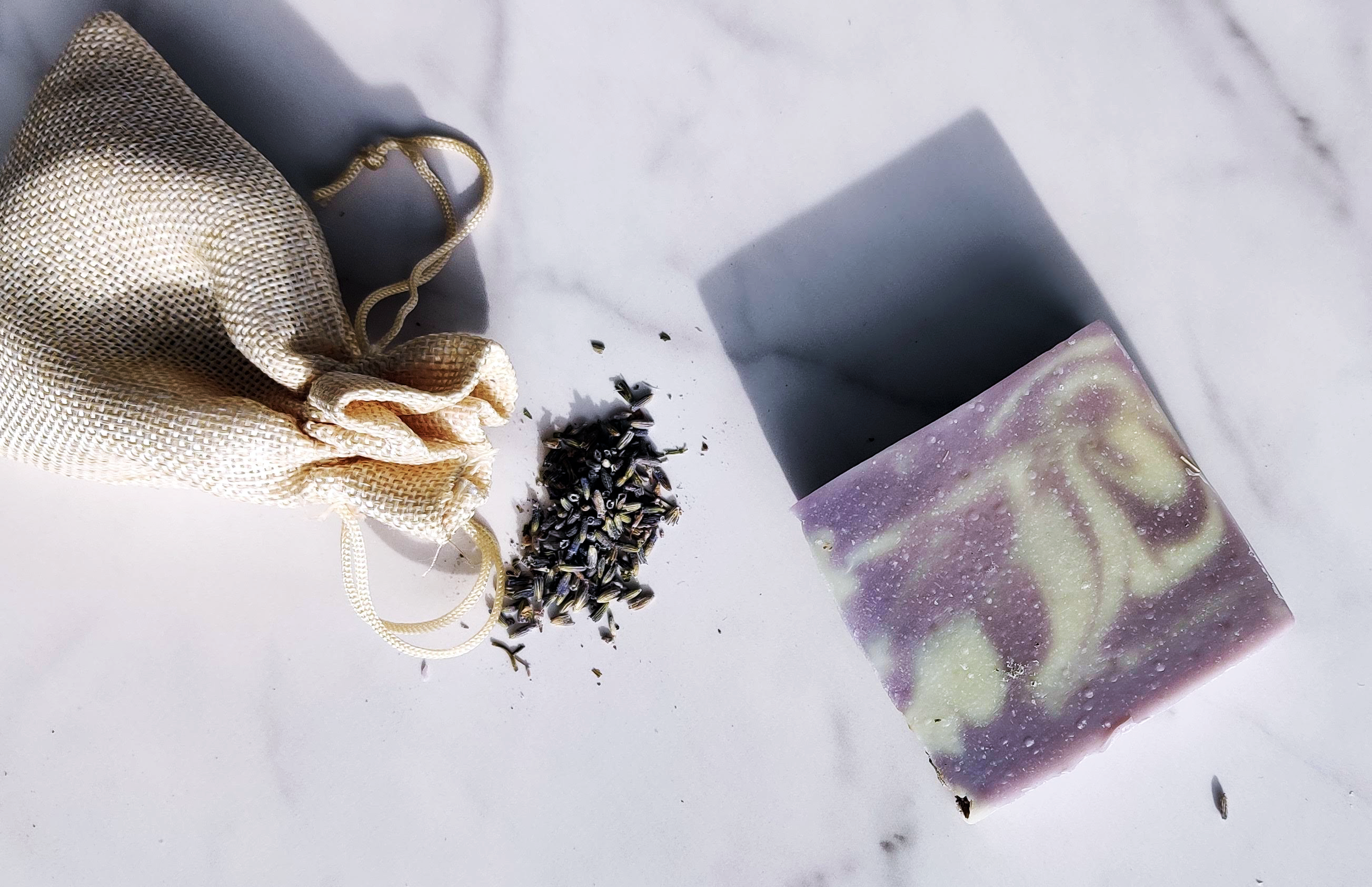 A beige mesh bag with a drawstring, some scattered lavender-colored seeds, and a purple and white marbled soap bar on a white surface.