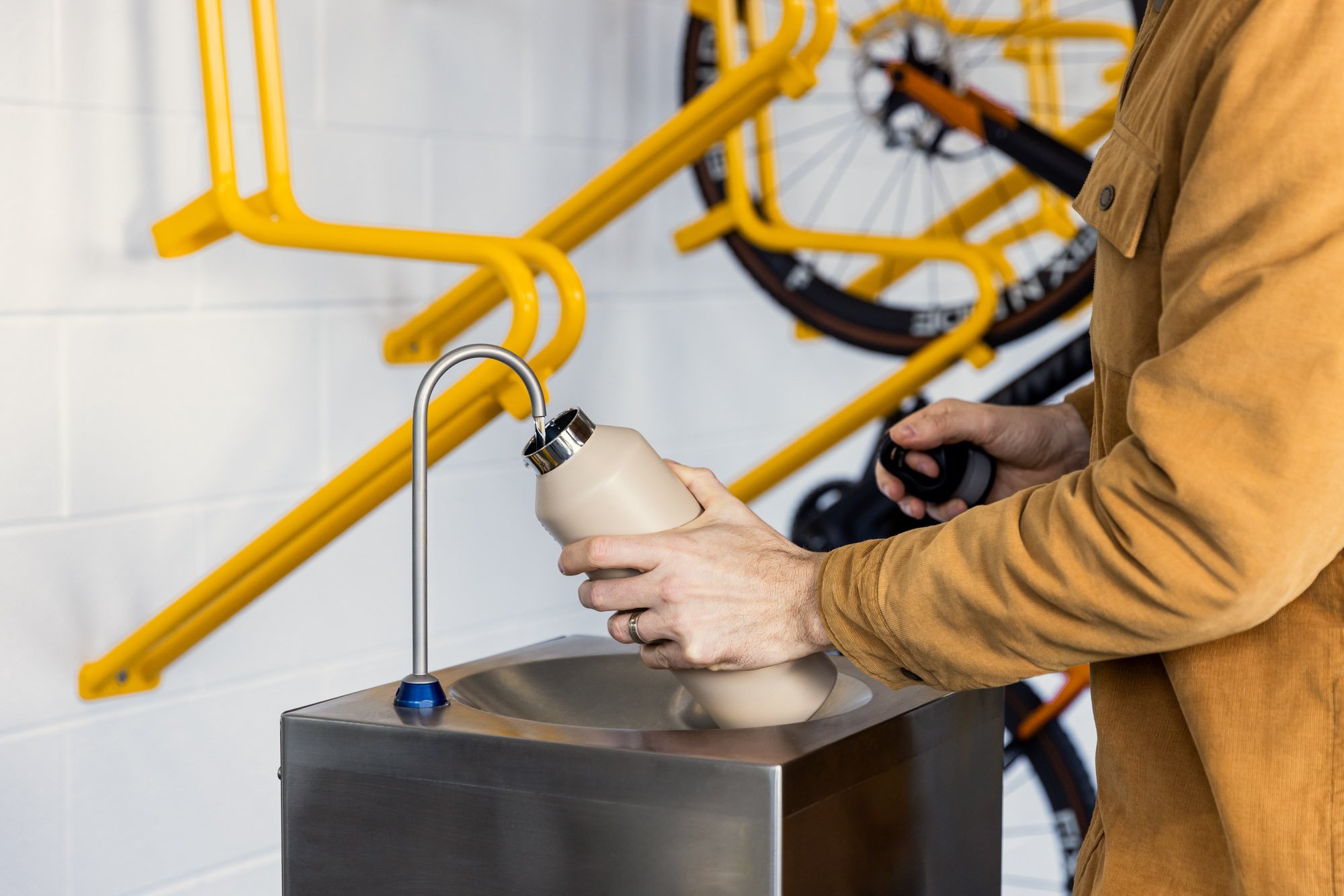 Person filling a water bottle at a water fountain in a bike shop, with yellow bikes hanging on the wall in the background.
