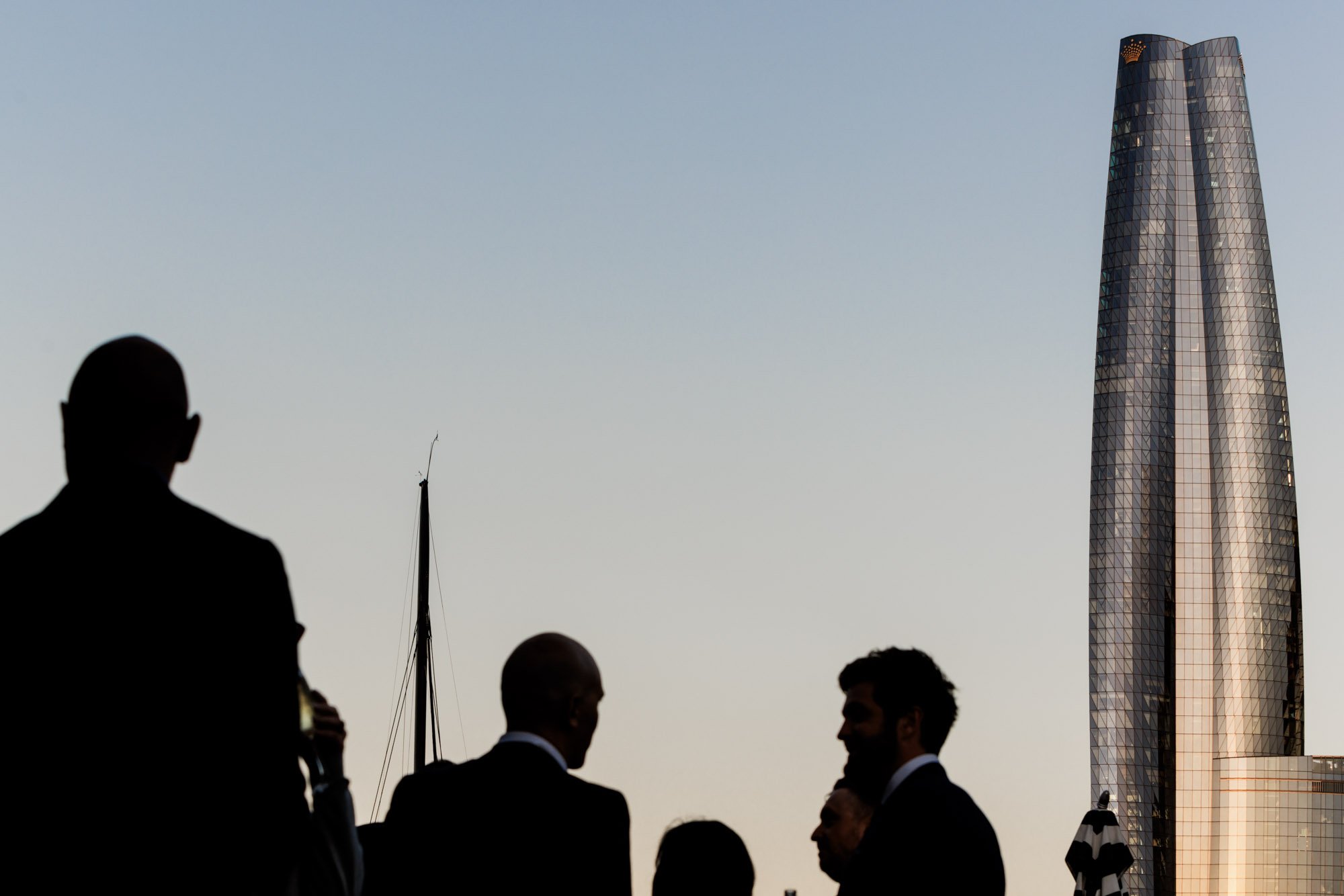 Silhouettes of people in formal suits talking outdoors with a modern glass skyscraper and a sailboat in the background during sunset.