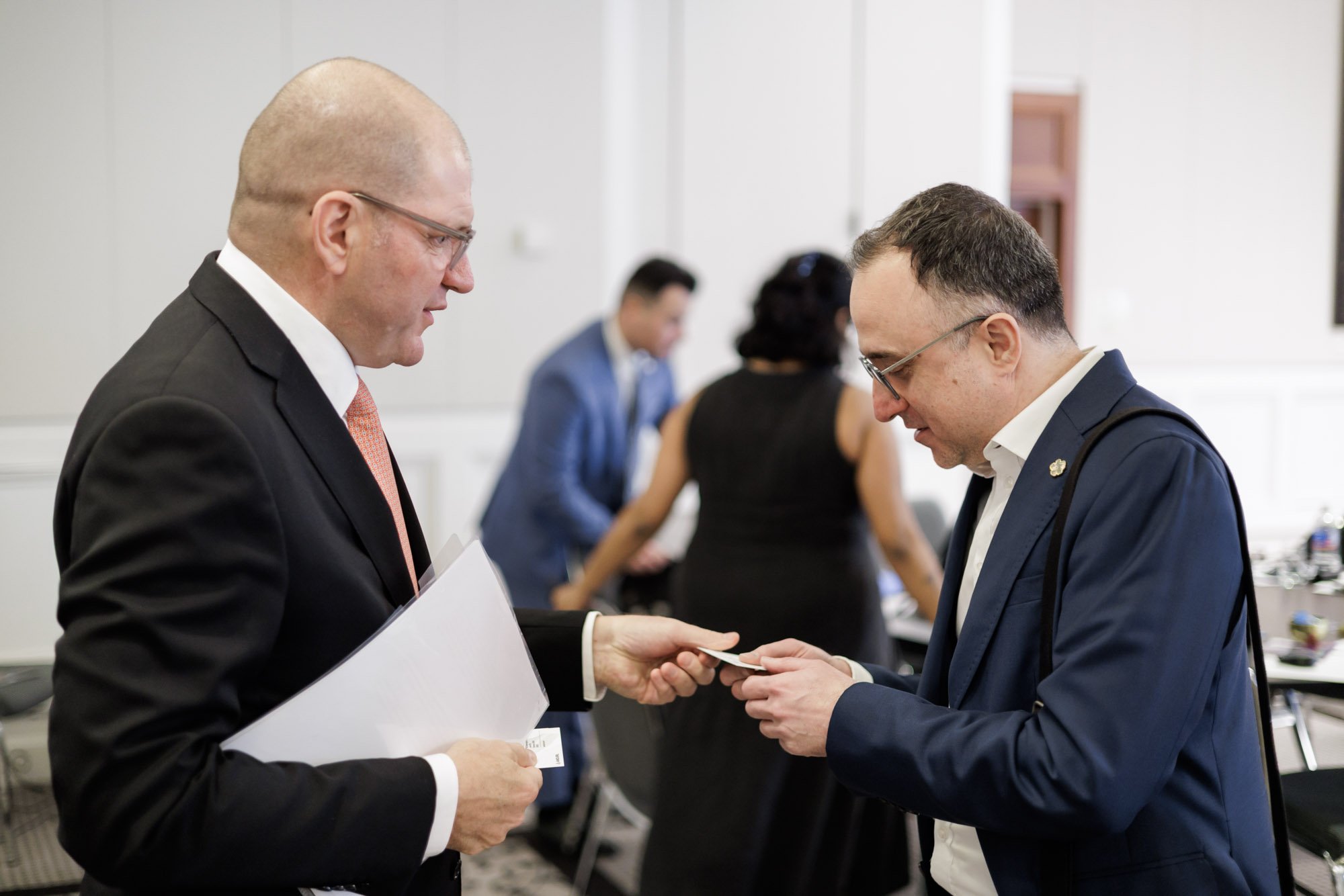 Two men in business suits exchanging business cards at a professional event, with other attendees in the background.