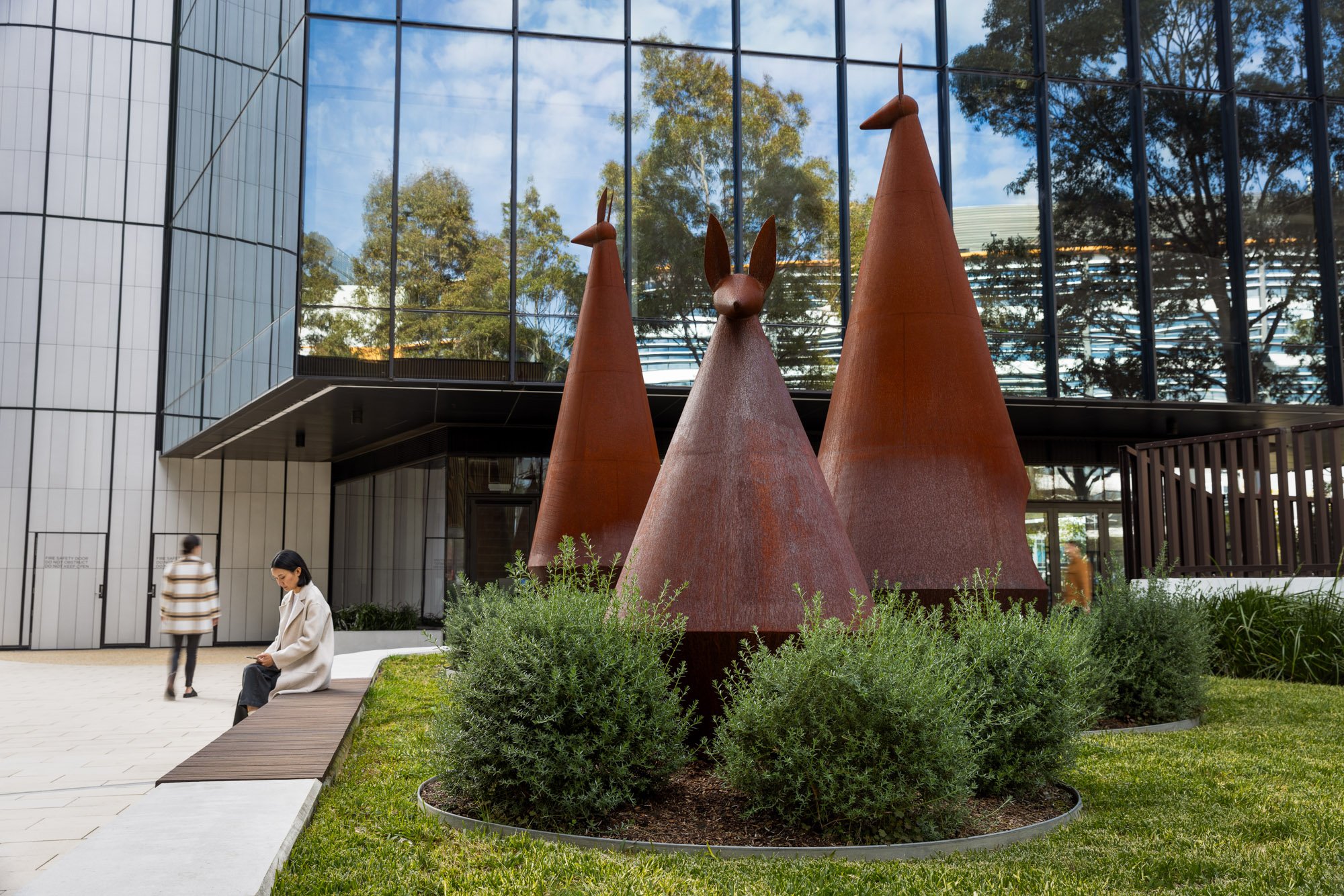 Modern glass building with three large rust-colored metal sculptures of stylized animals, resembling kangaroos or wallabies, surrounded by green bushes and two women sitting and walking nearby.