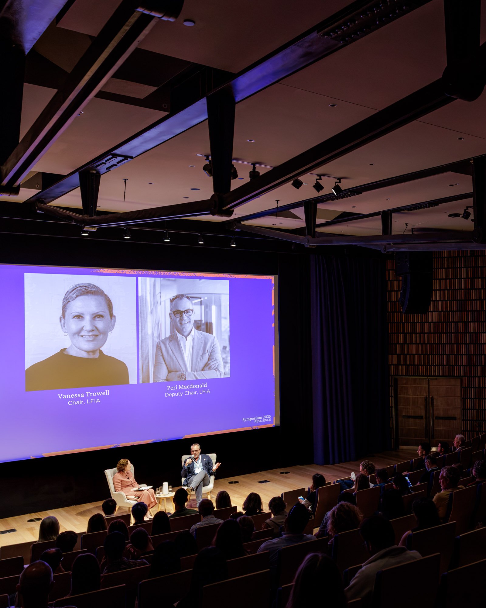 A conference room with a stage and a large screen displaying two speaker portraits and their names, Vanessa Trowell and Peri Macdonald, during a symposium on resilience. The audience is seated facing the stage, and there are three people on the stage: a woman in a peach-colored dress, a man with glasses and a suit, and another man speaking into a microphone.