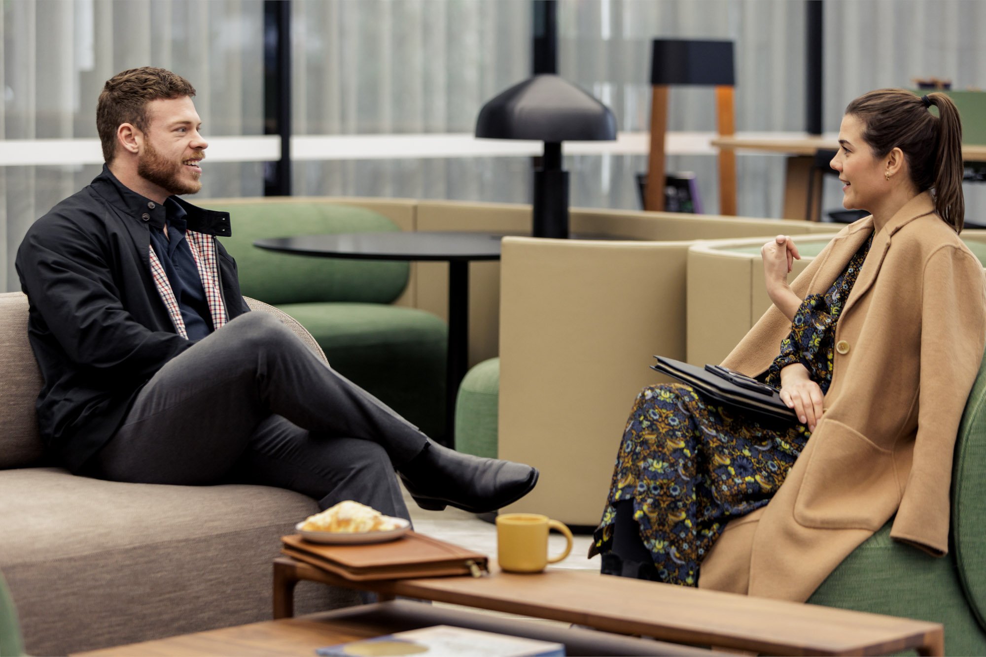 A man and a woman are sitting on sofas in a modern, open office space, engaged in a conversation.