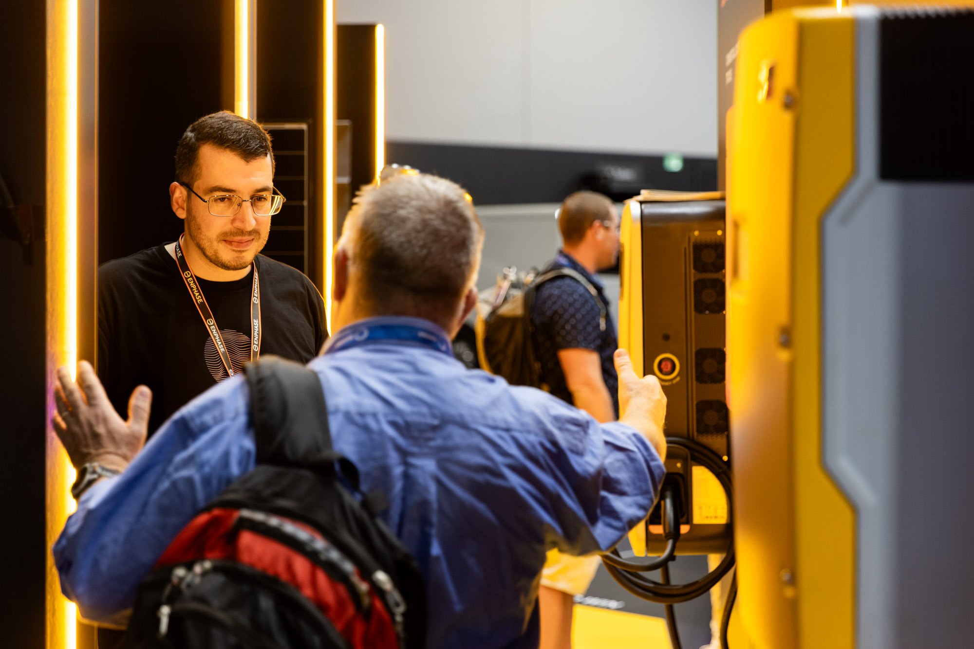 Two men engaged in conversation at a booth, with electronic equipment and another person in the background at a technology event or conference.