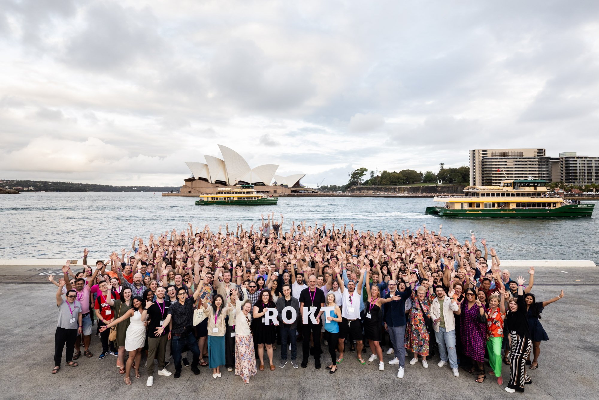 Large group of people gathered outdoors near a body of water with the Sydney Opera House and boats in the background, holding up letters spelling RCOT.
