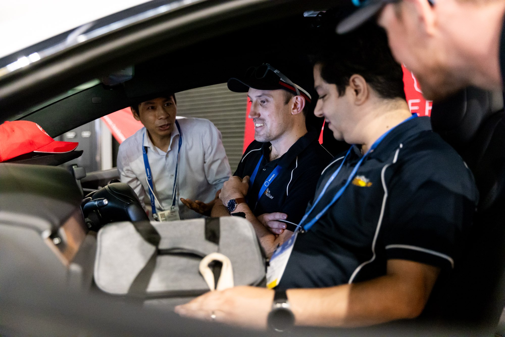 Four men sitting inside a car having a discussion, with one man standing outside the car. The men appear to be in a professional setting, possibly related to racing or automotive industry, with one wearing a cap and another wearing sunglasses on his 