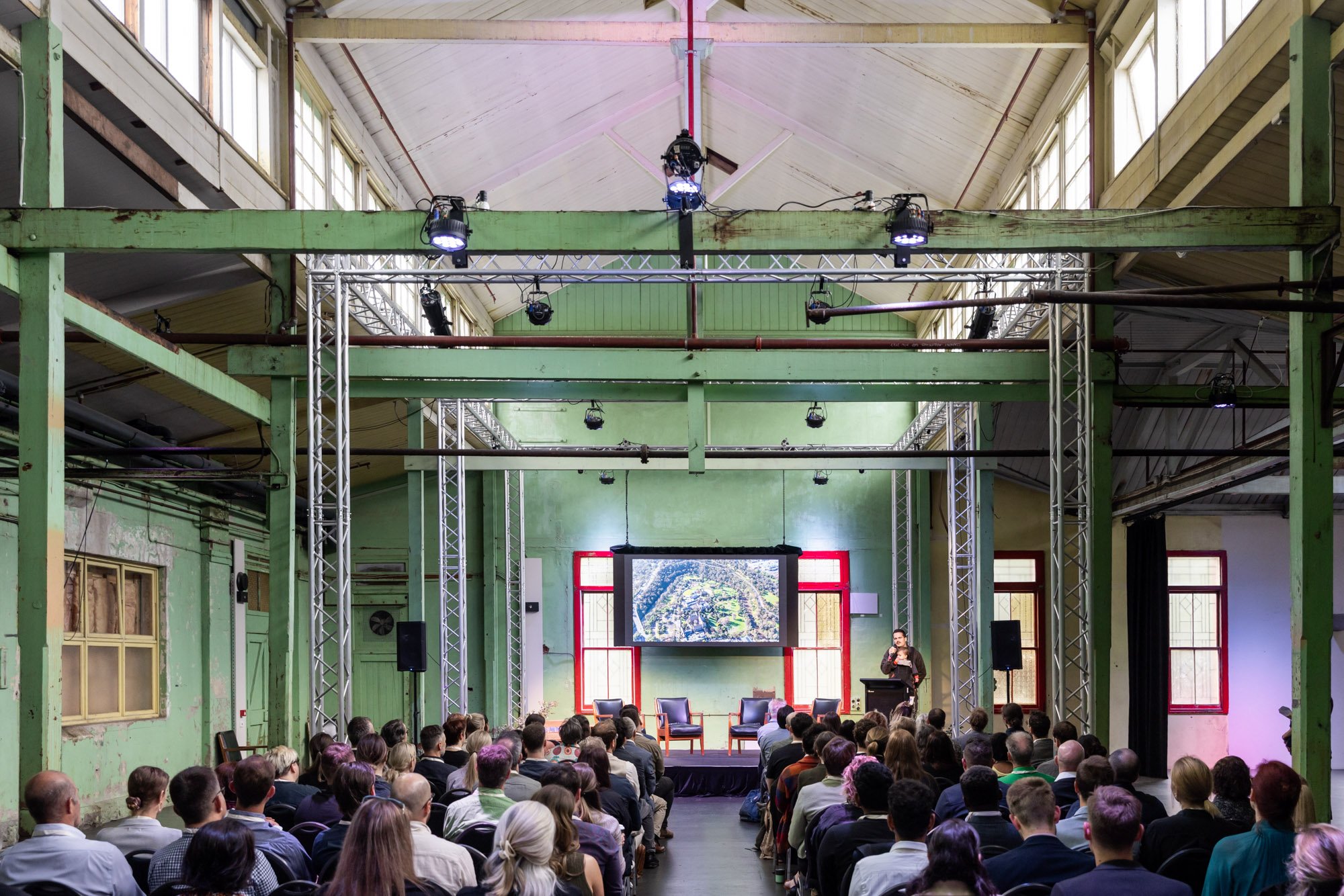 A conference or presentation event taking place in an industrial-style venue with green painted walls and large windows. A speaker is on stage near a large screen displaying an aerial view of a landscape. The audience is seated facing the stage, with