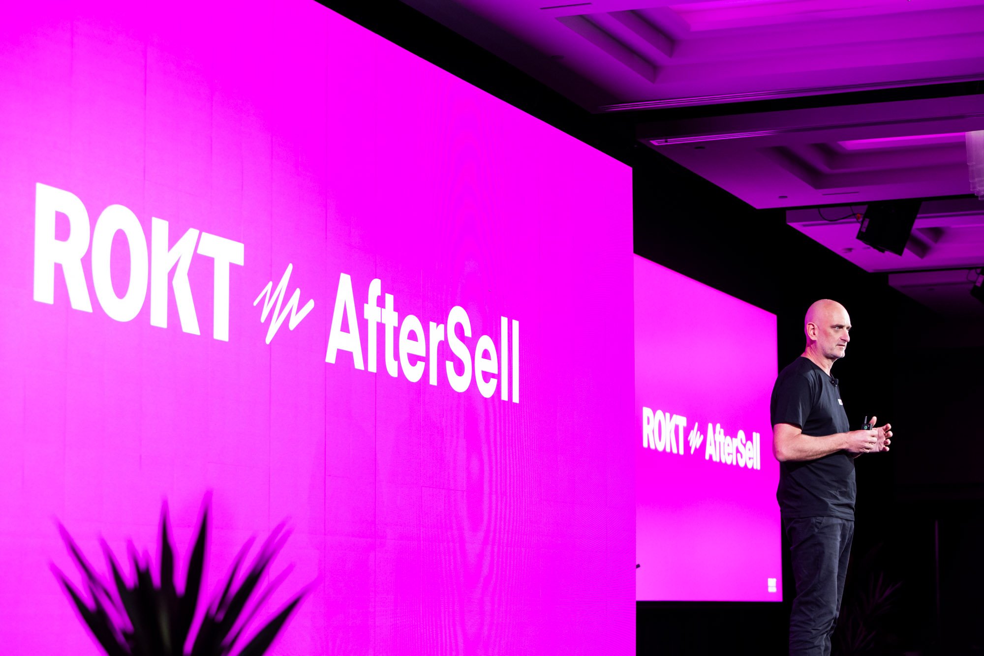 A man standing on stage with a microphone in hand, against a large pink screen displaying 'ROKT w/ AfterSell' in white text. The room appears to be a conference or presentation setting.