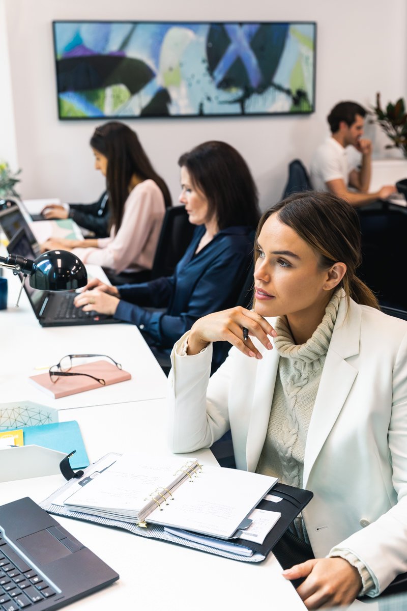 Business meeting or conference with women and men working at desks in an office, some using laptops and others writing, with a large colorful screen on the wall.