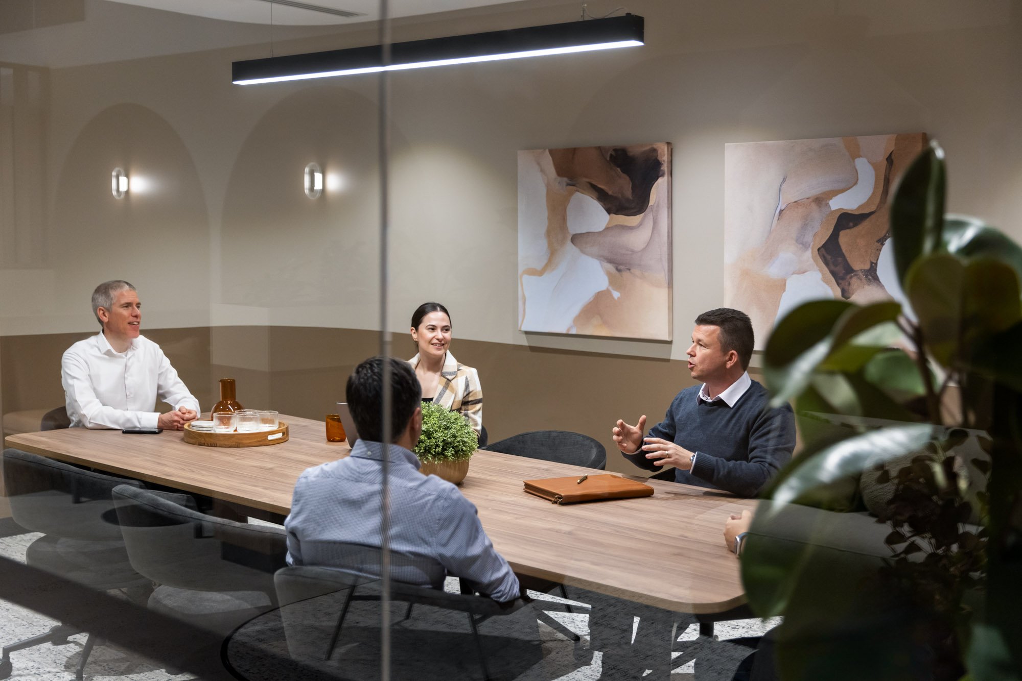 Four people are sitting around a wooden conference table in a modern office, engaged in a discussion. A man on the right is speaking, while others listen. The room has beige walls, interactive abstract art, and is softly lit.