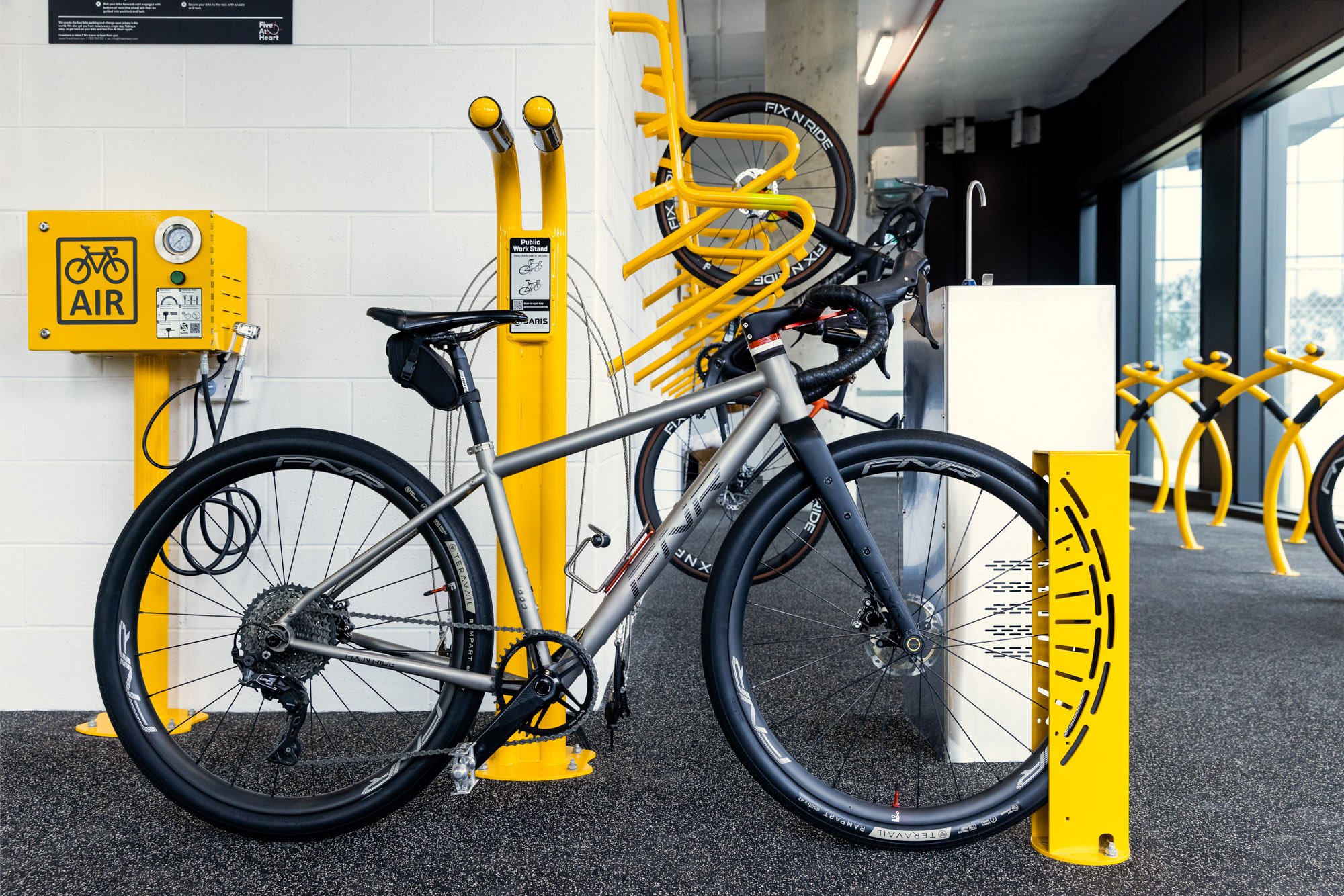 Indoor bike storage area with yellow bike racks, a parked silver and black bicycle, and a yellow bike air pump station on a white wall.