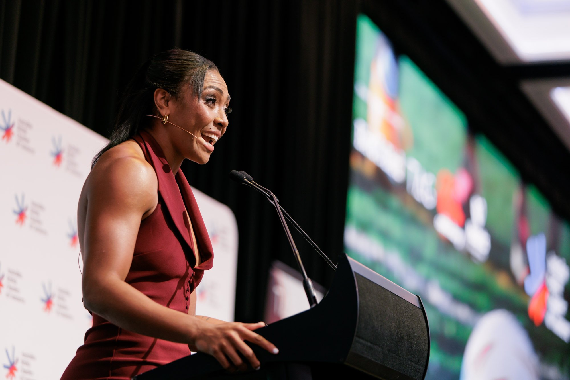 A woman standing at a podium speaking at a conference with a microphone headset and a large screen in the background.