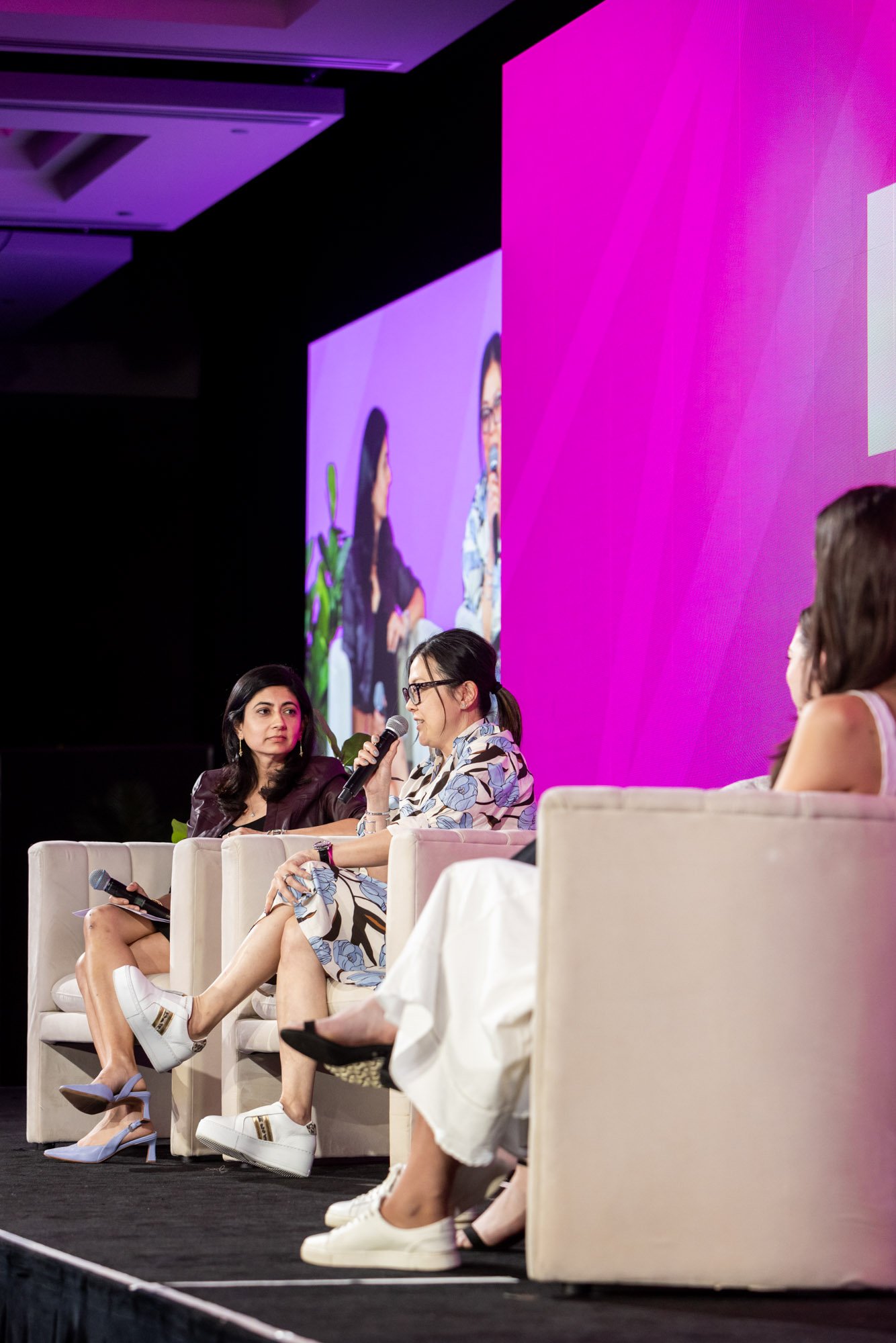 Women participating in a panel discussion on stage at an event, sitting in light-colored armchairs with a large screen behind them displaying a close-up of the women.