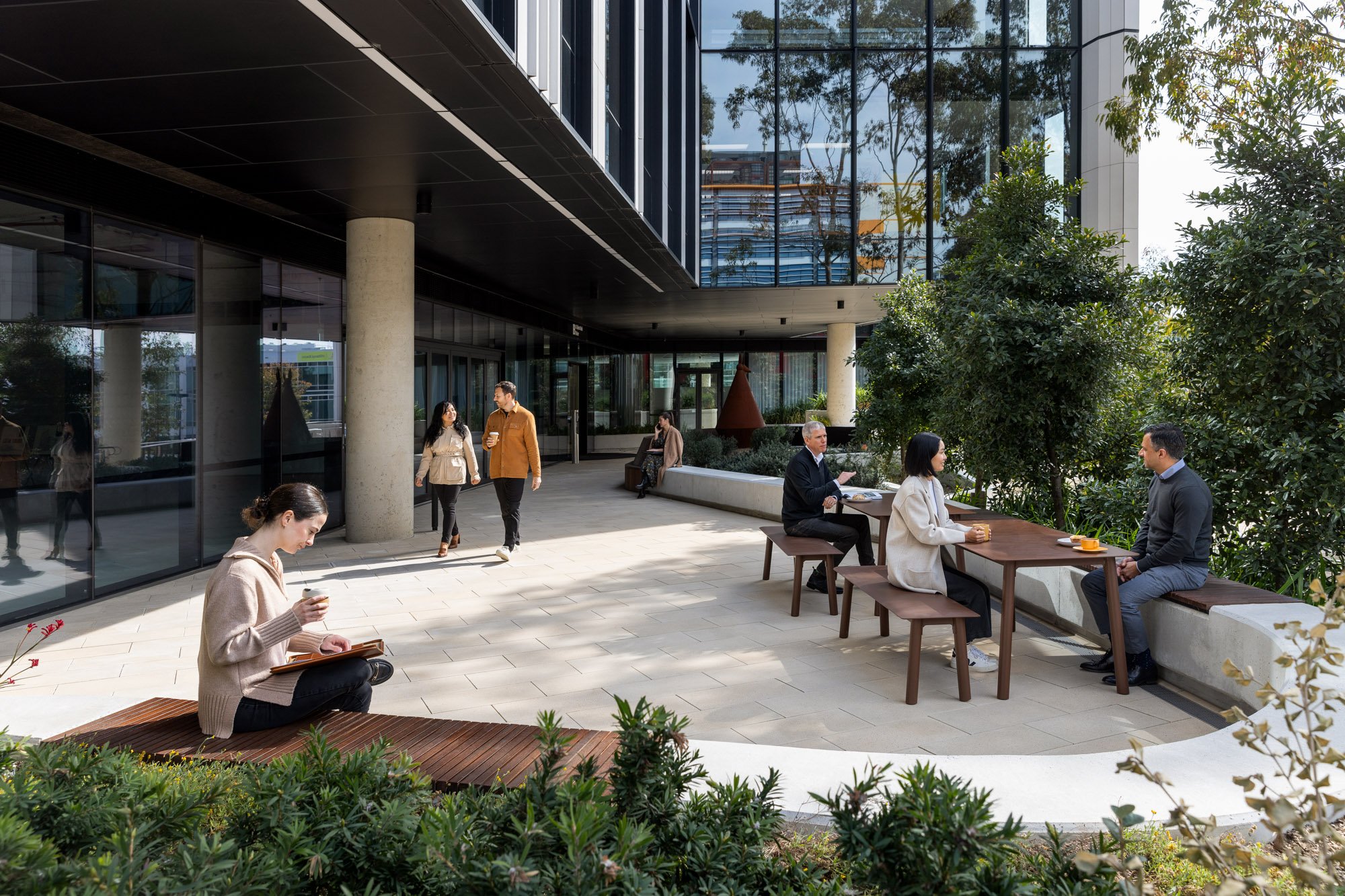 People sitting and chatting at an outdoor patio of a modern building with greenery and trees.