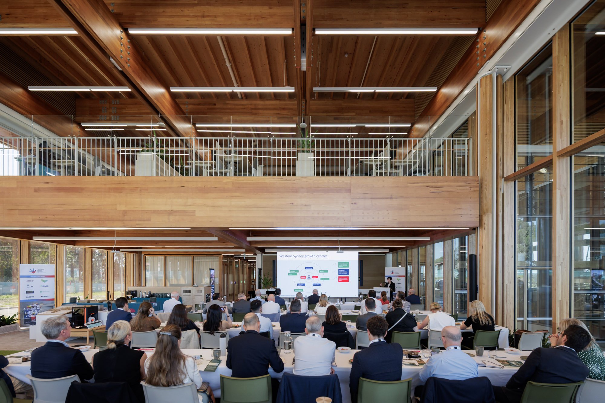 Business conference in a modern wooden-paneled conference room with attendees seated facing a large presentation screen displaying a diagram. There are two levels, with the upper level having chairs and tables. Large windows on the sides allow natura