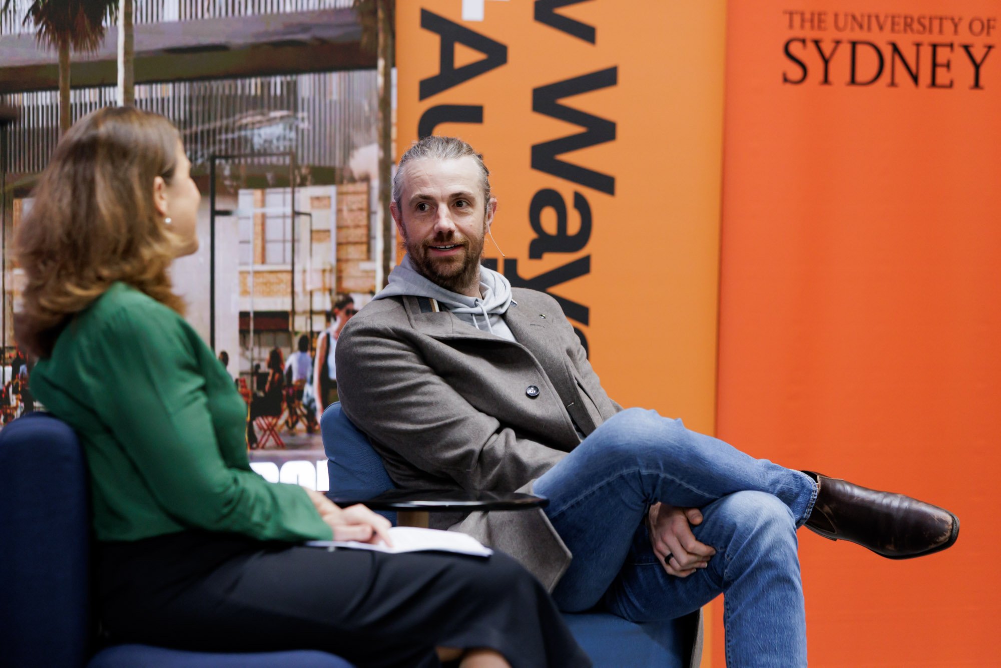 A man and a woman seated on chairs, engaged in conversation at an event related to the University of Sydney, with a backdrop displaying part of the university's name and campus scene.