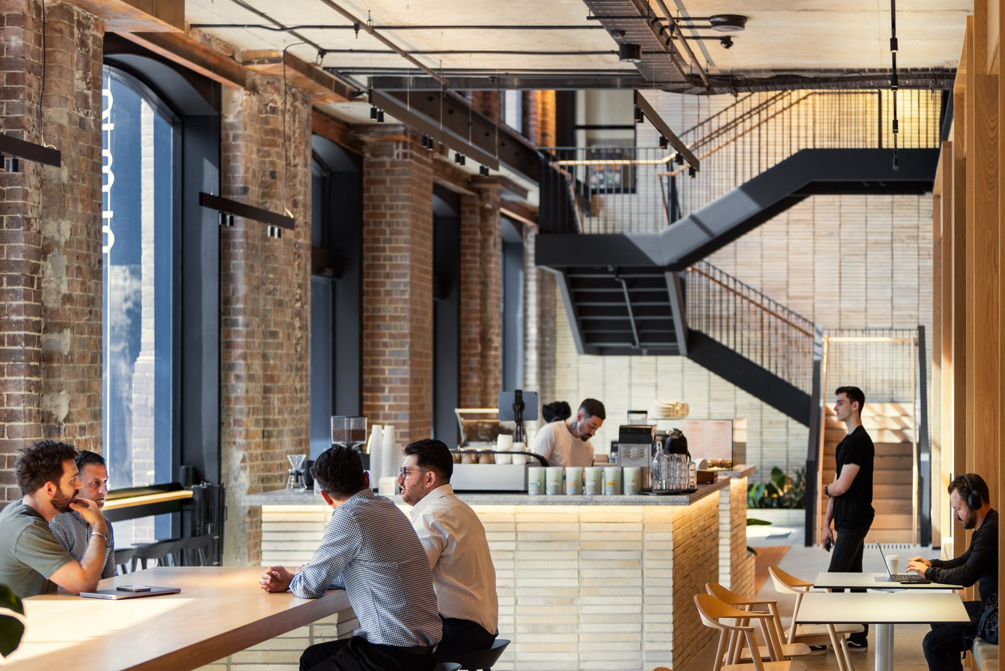 Interior of a modern coffee shop with a brick wall, large windows, and industrial design elements. Four people are seated at a high wooden table, and a barista is preparing drinks behind the counter. A woman is working on a laptop at a small table, w