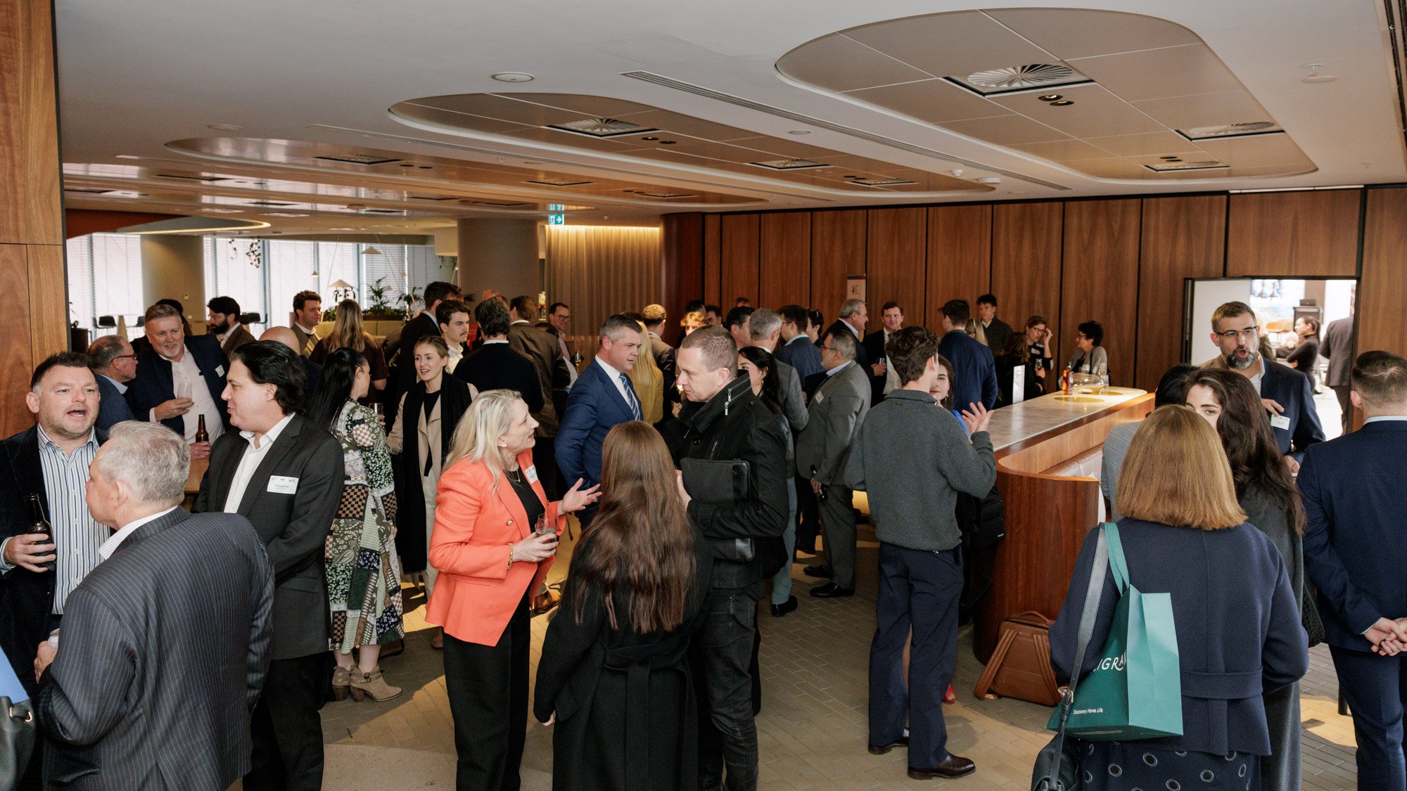 Crowd of professionally dressed people socializing at a conference or networking event in a modern, wood-paneled indoor space.
