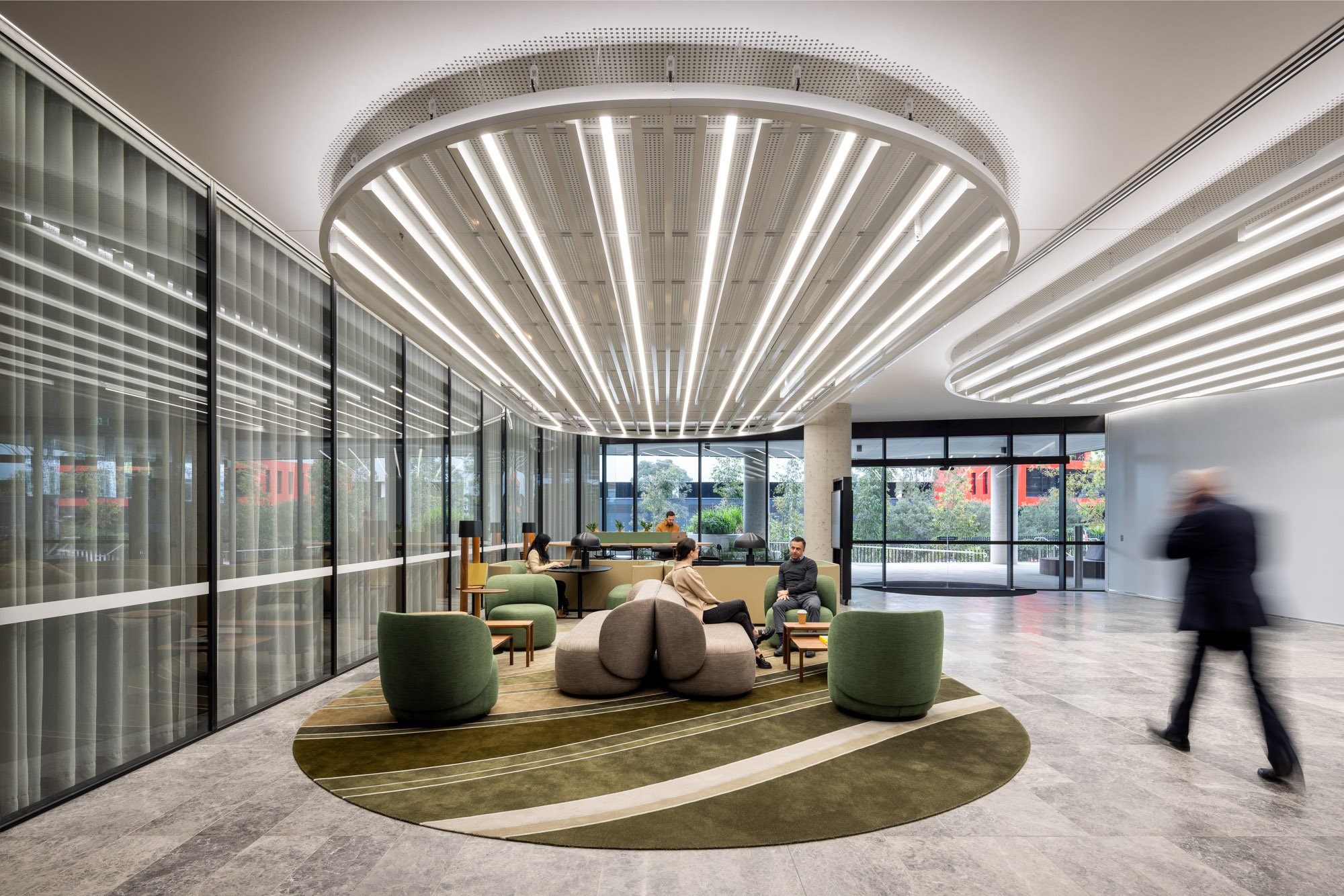 Modern lobby with green and beige seating, large glass walls, curved ceiling with integrated lighting, and a man walking past.