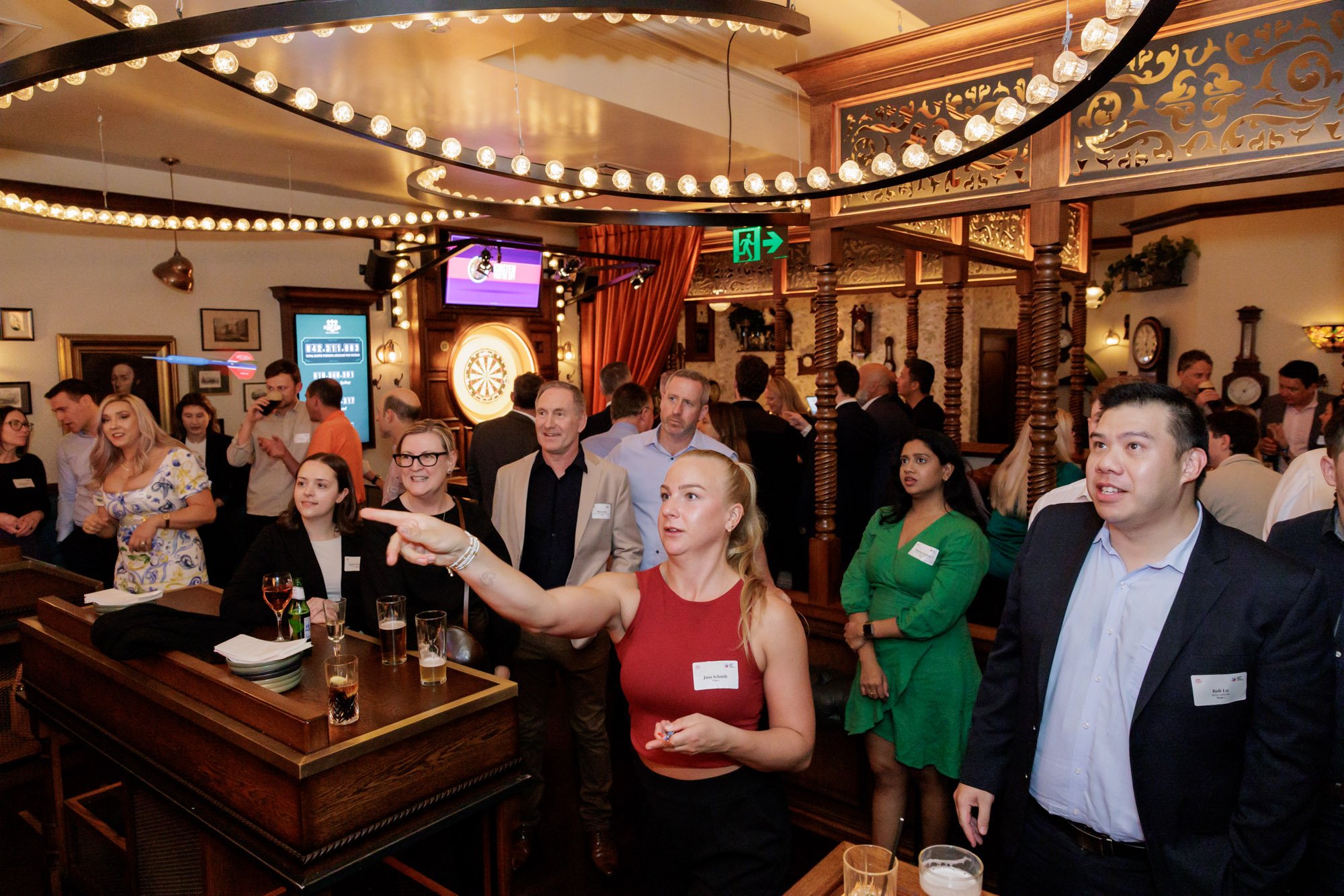 People socializing in a bar or restaurant with ambient lighting, dartboard, and decorative wood paneling, some standing and talking, others watching a woman in a red top pointing.