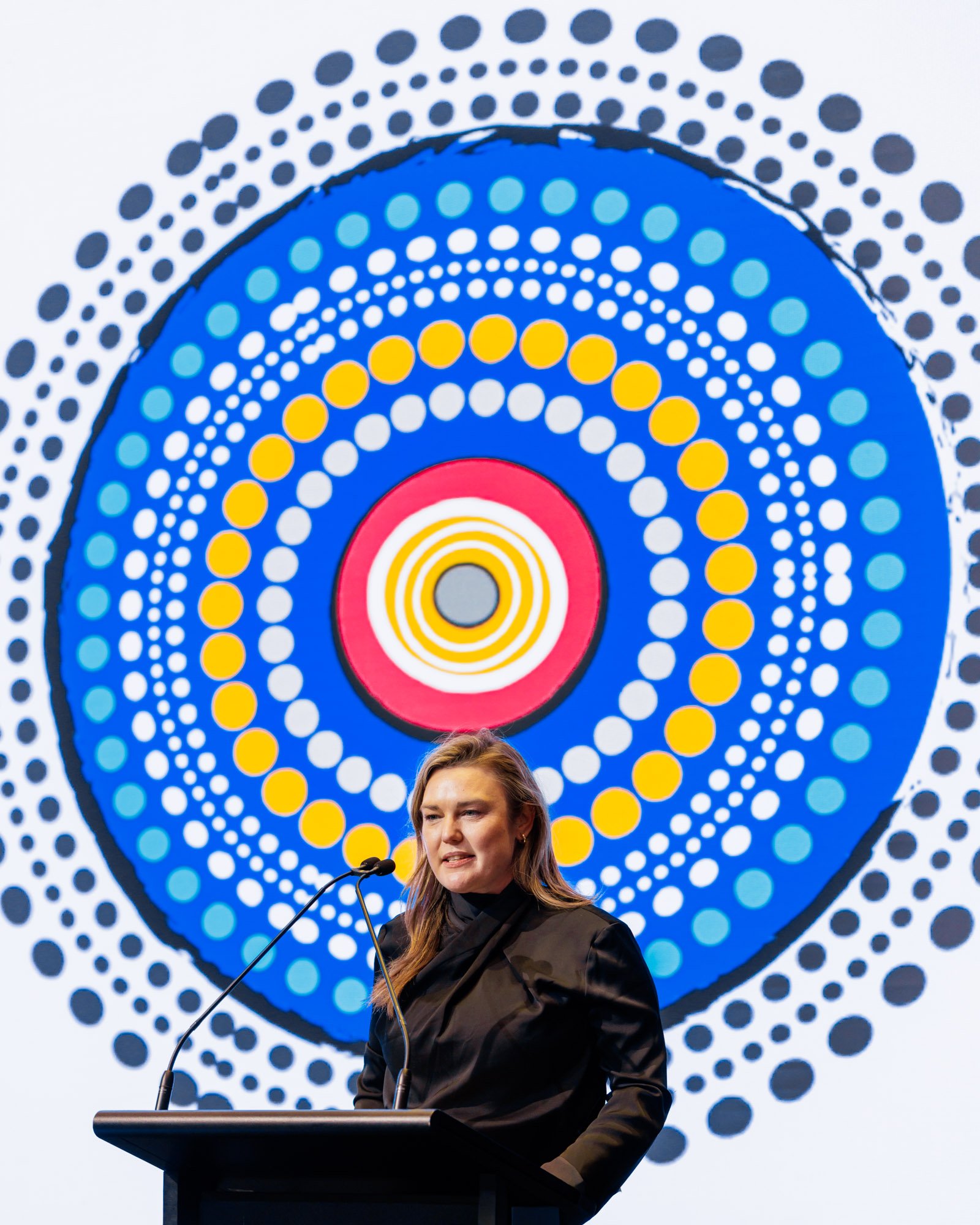 A woman speaking at a podium at a conference with a microphone, standing in front of a large, colorful, circular, dotted graphic design in the background.