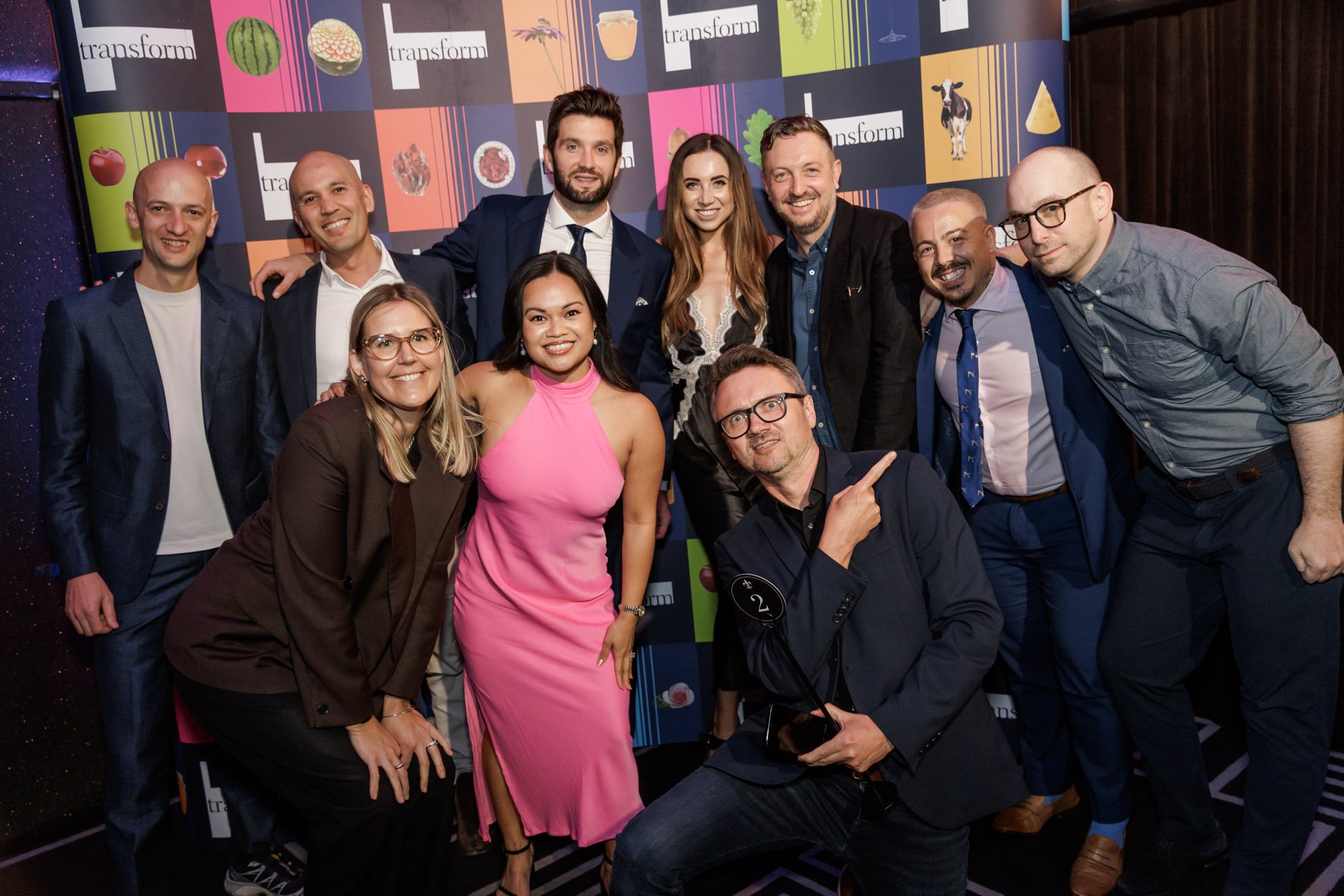 Group of eleven people posing together at an event, in front of a colorful backdrop with the word 'transform' and images of various fruits and vegetables.