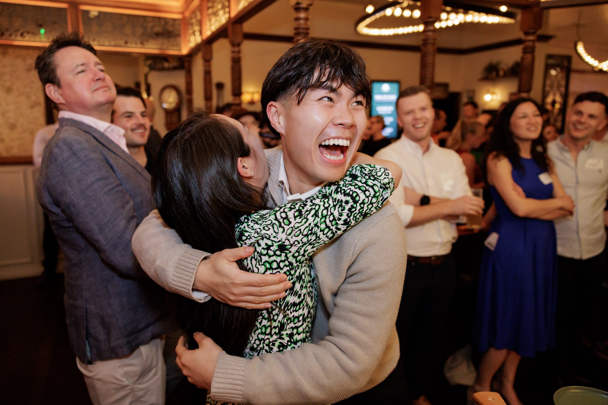 People at a social gathering, with a young man and woman hugging and smiling in the foreground, surrounded by others who are smiling and watching in a warmly lit room.