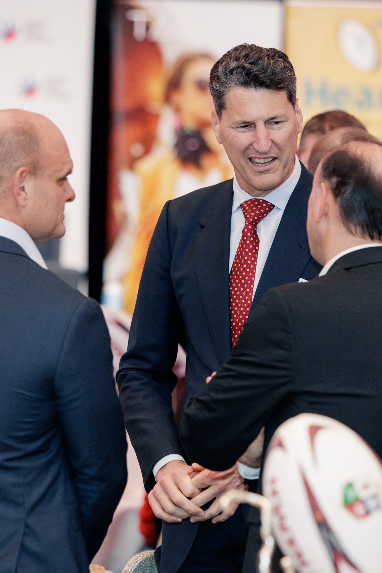 Three men in suits engaged in a conversation at a professional event, with one man in the center smiling and making a gesture with his hands.